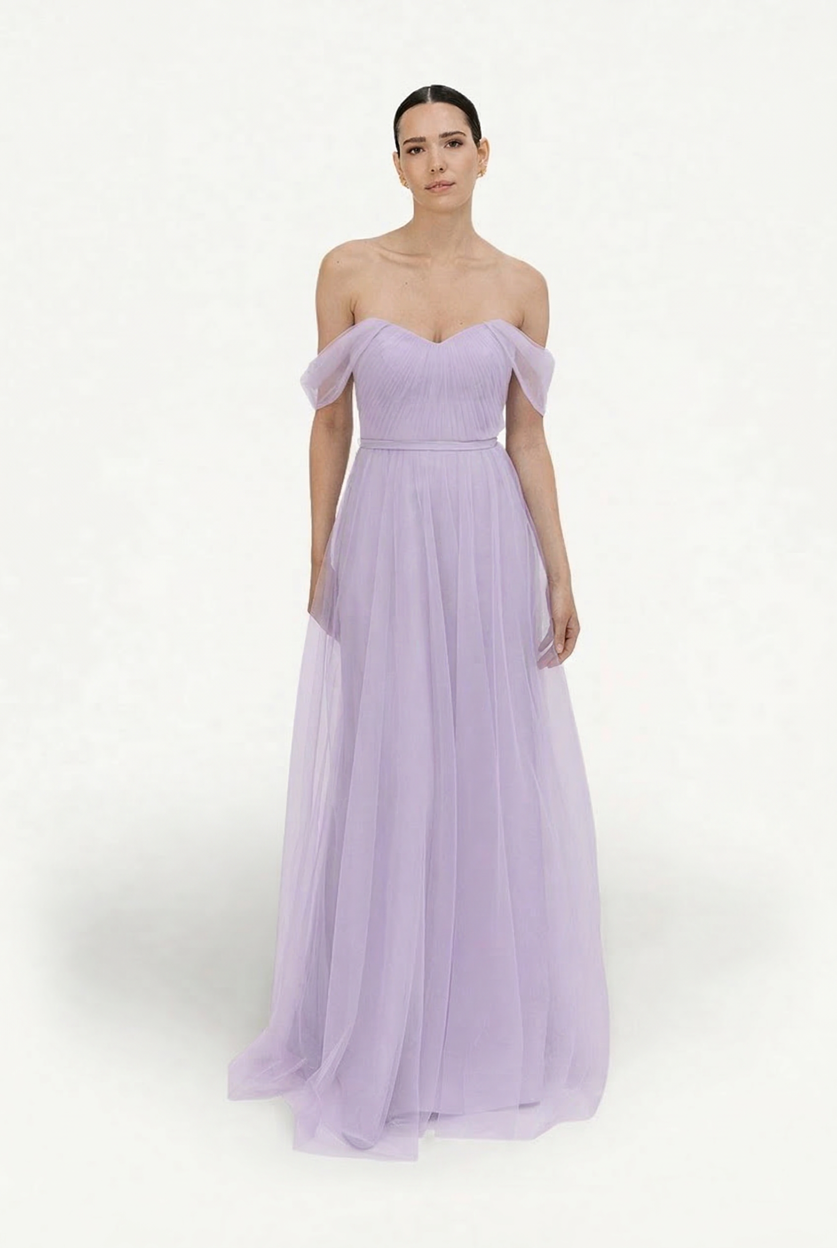 A woman stands against a plain white background, wearing the TH&TH Bardot Bridesmaid Dress in Lilac Mist Tulle with an off-shoulder design, fitted bodice, and flowing floor-length skirt for an elegant look.