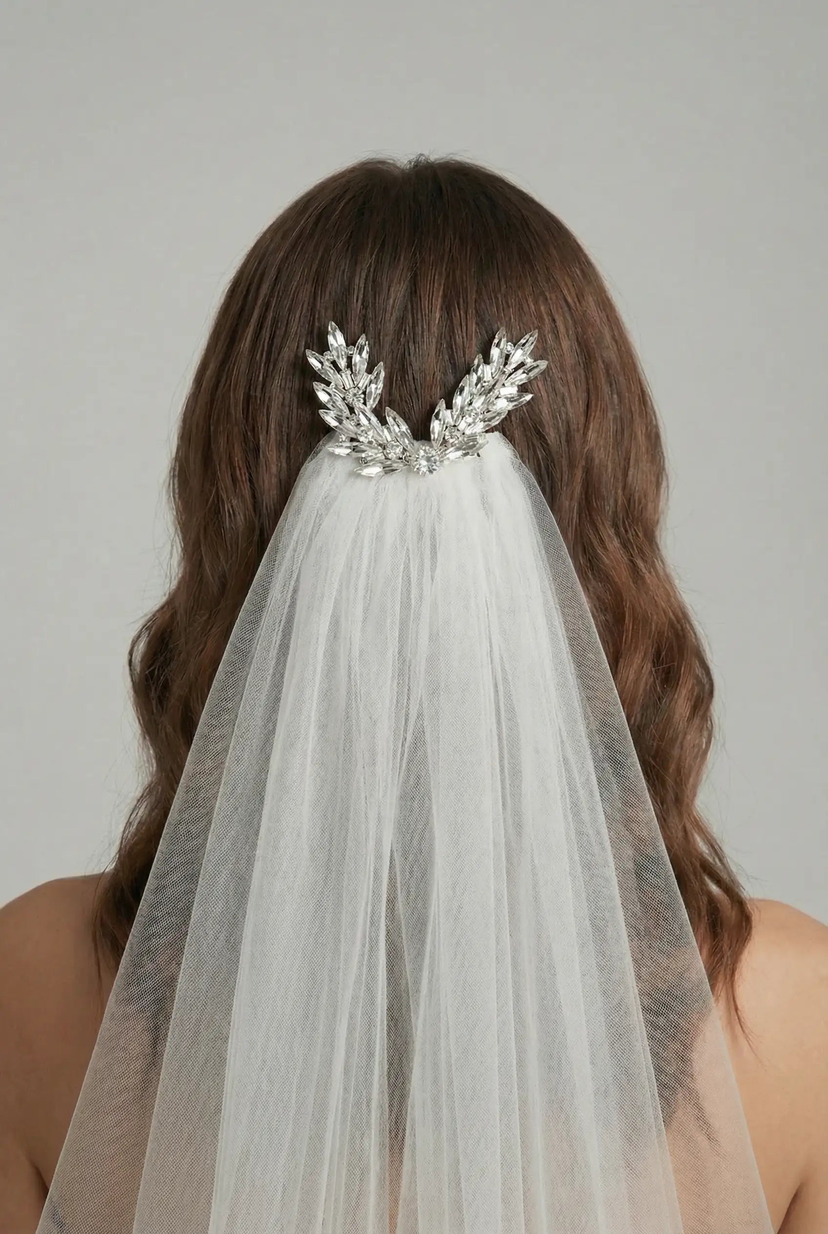 A woman with wavy brown hair is shown from behind, wearing the TH&TH Beaumont Crystal Leaf Hair Comb, an elegant wedding accessory that adds sparkle to her bridal look beneath a long white veil.