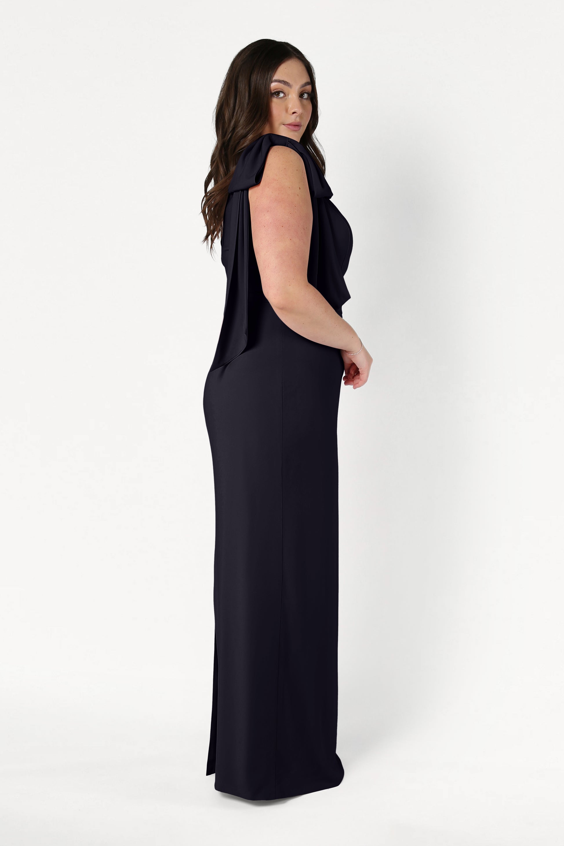 A woman with long brown hair stands in profile against a plain white background, looking over her shoulder and showcasing the Valentina Bridesmaid Dress in Black by TH&TH.
