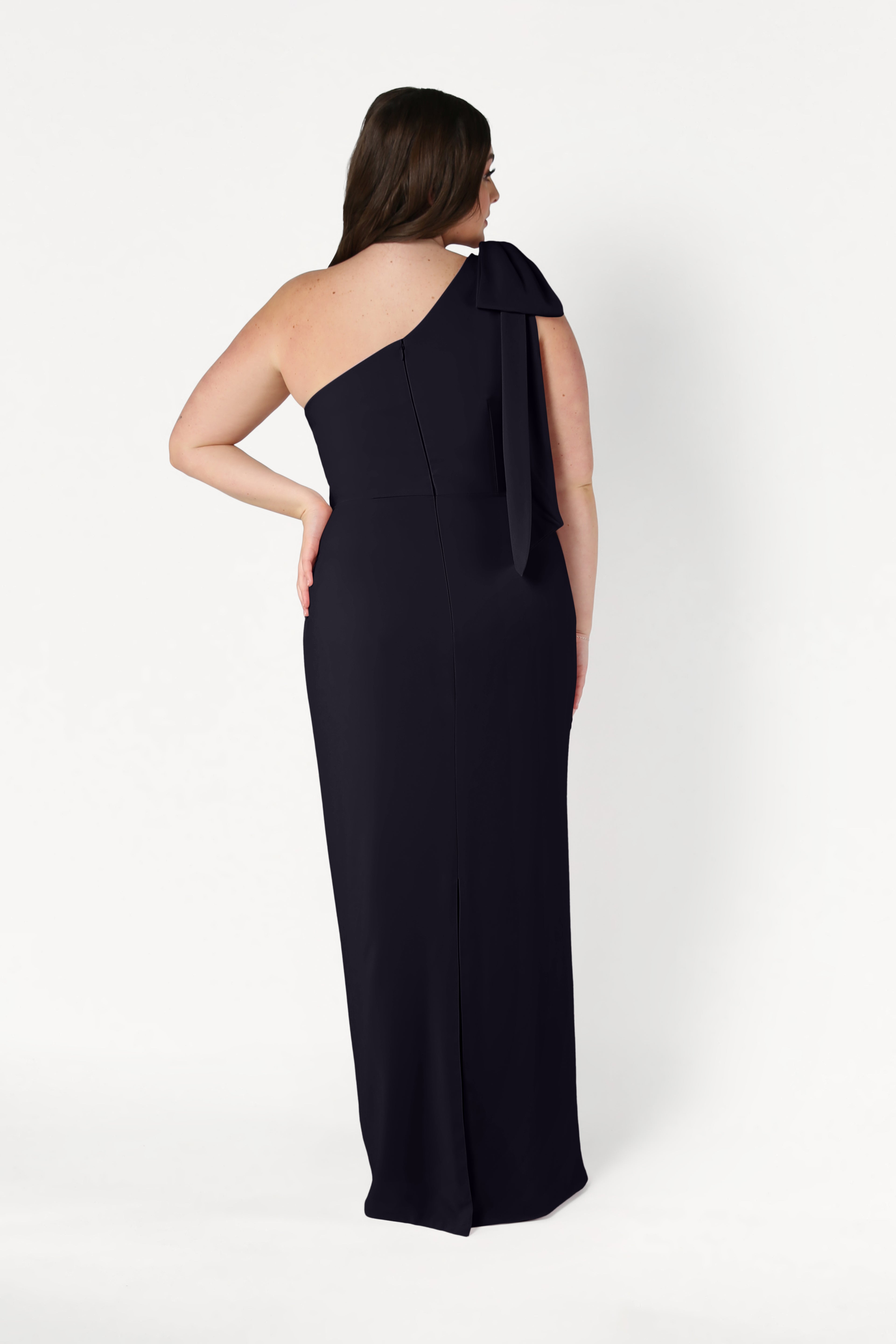 A woman faces away, wearing the TH&TH Valentina Bridesmaid Dress in Black—a long, fitted, one-shoulder gown with a bow detail on the shoulder—set against a plain white background.