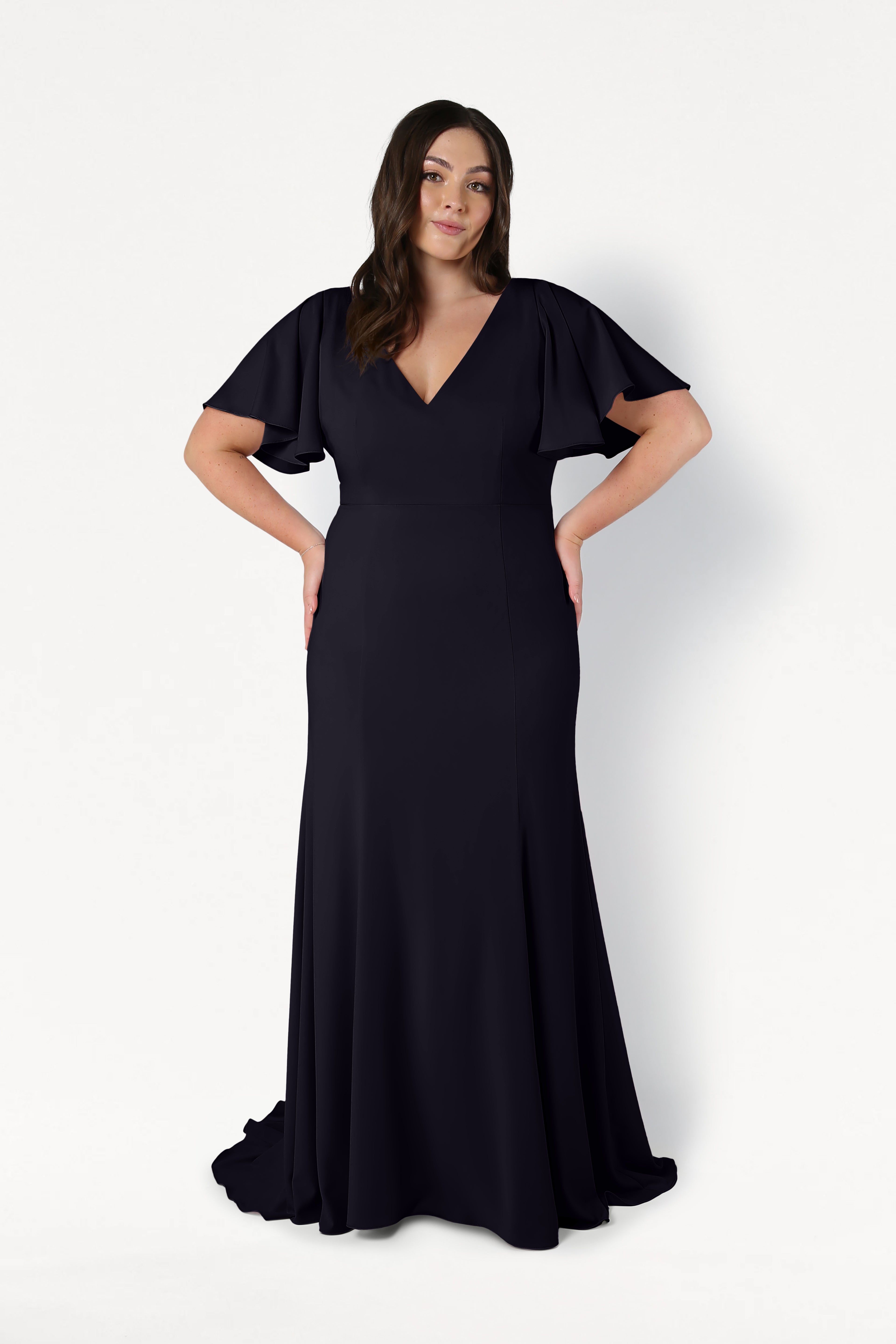 A woman poses against a white background wearing the TH&TH Celeste Moss Crepe Bridesmaid Dress in Black. The floor-length gown features flutter sleeves and a V-neckline. Her long brown hair falls in loose waves as she rests her hands on her hips.