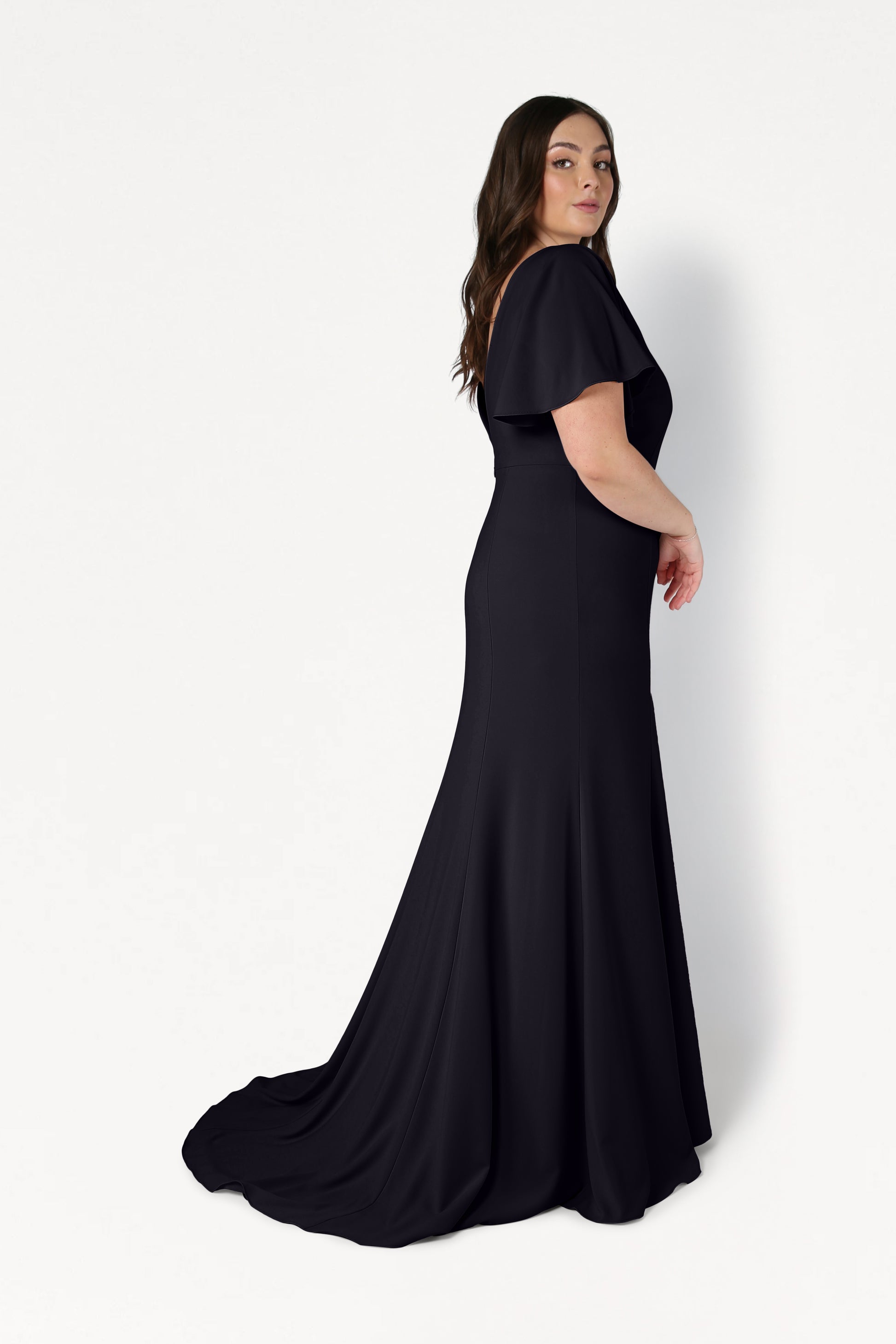 A woman with long brown hair models the TH&TH Celeste Moss Crepe Bridesmaid Dress in Black, a fitted floor-length gown with short flutter sleeves, posing sideways against a plain white background.