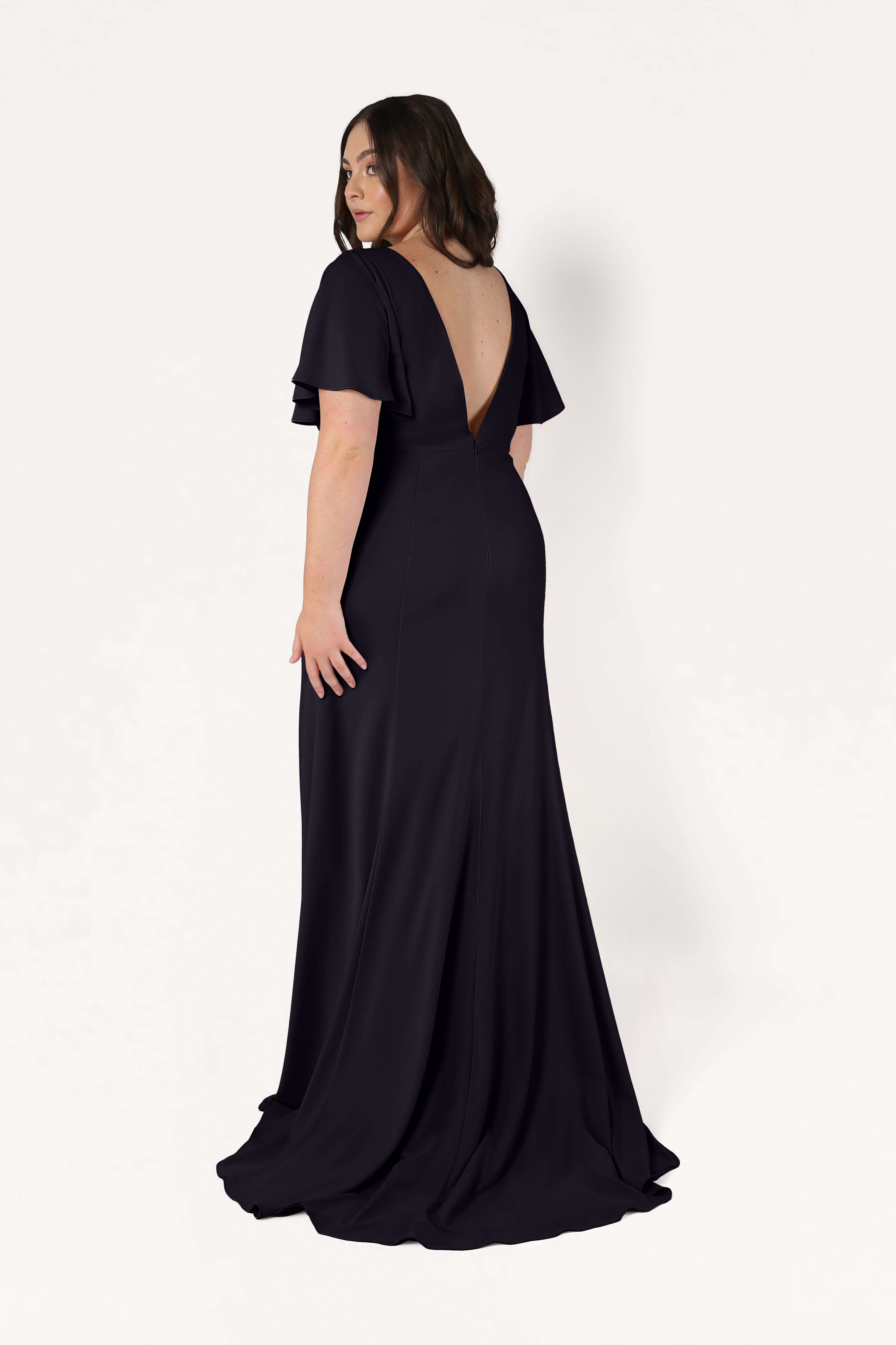 A woman with shoulder-length brown hair models the floor-length Celeste Moss Crepe Bridesmaid Dress in Black by TH&TH. She stands on a white background, facing away and looking over her left shoulder to reveal the deep V-back.