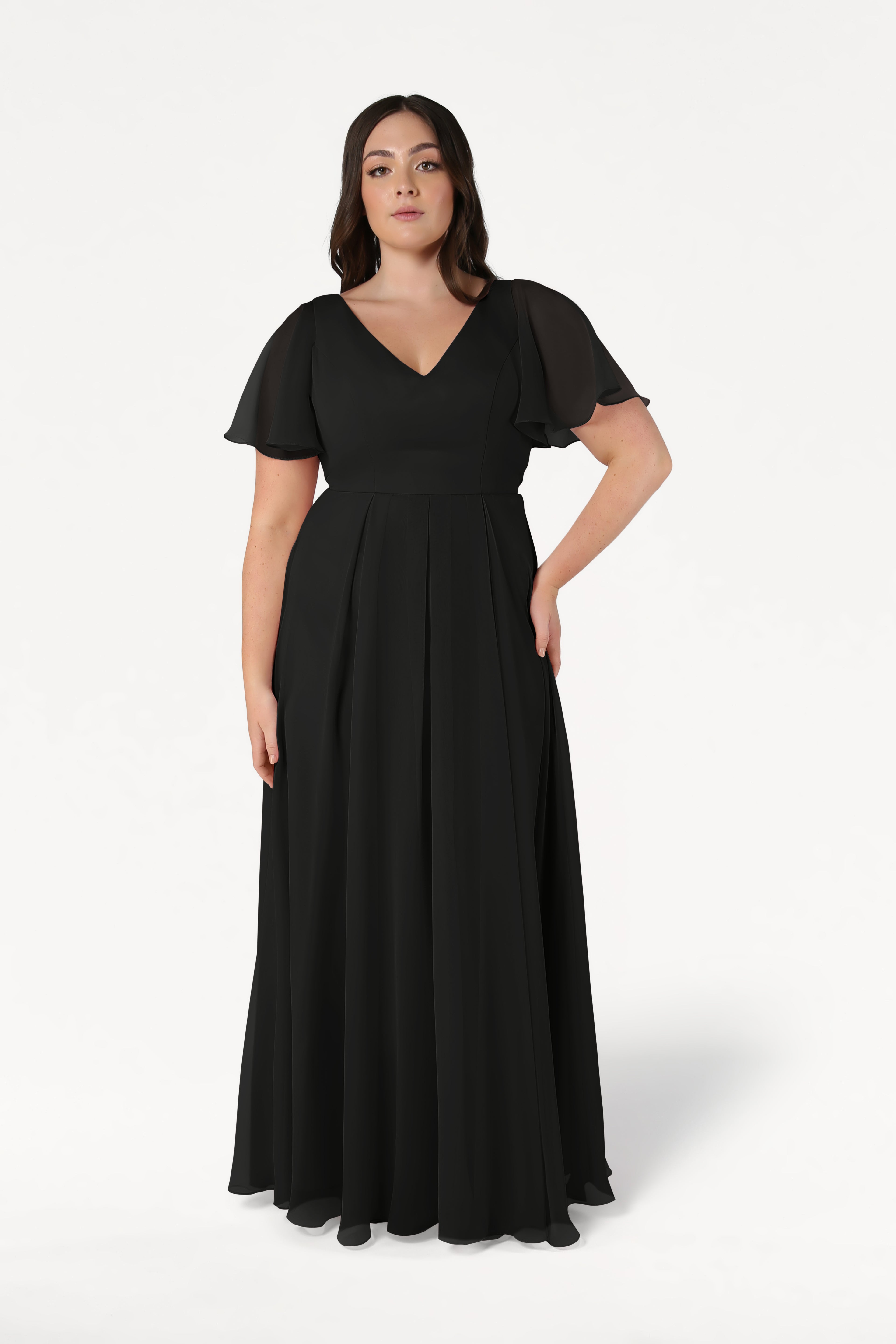 A woman stands against a plain white background wearing the TH&TH Phoebe Bridesmaid Dress in black chiffon, featuring a floor-length skirt, short sleeves, and a V-neckline.