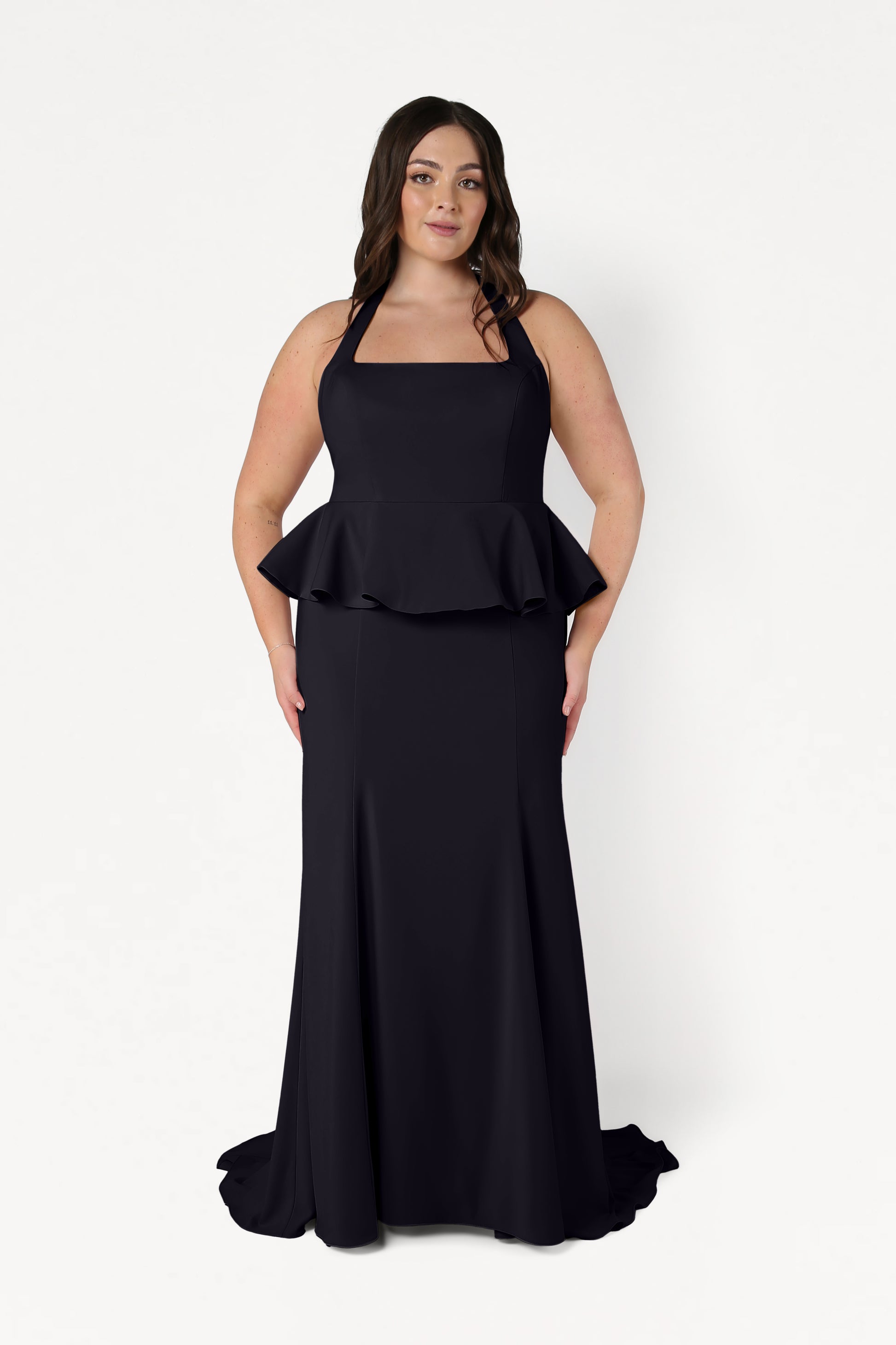 A woman with long dark hair wears the TH&TH Peony Bridesmaid Dress in Black—a size-inclusive, floor-length, sleeveless gown with a peplum waist—standing against a plain white background.