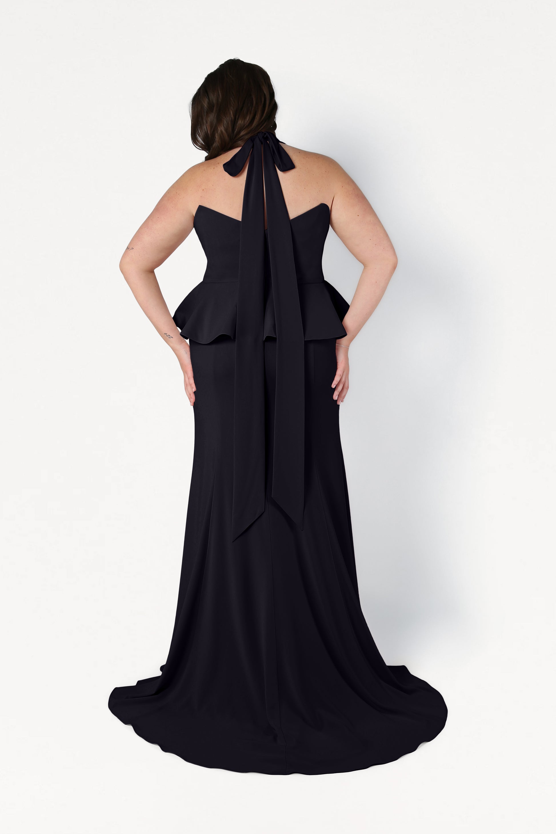 A woman faces away in the TH&TH Peony Bridesmaid Dress in Black—a size-inclusive, modern gown with a peplum waist, halter neck bow, and slight train—standing against a plain white background.