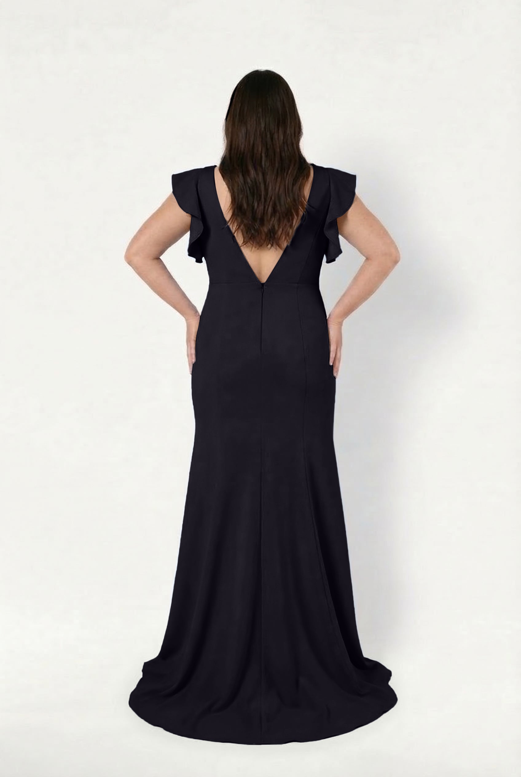 A woman with long brown hair stands facing away, wearing the TH&TH Cecelia Moss Crepe Bridesmaid Dress in Black, featuring a floor-length silhouette, deep V-back, and ruffled cap sleeves against a plain white background.