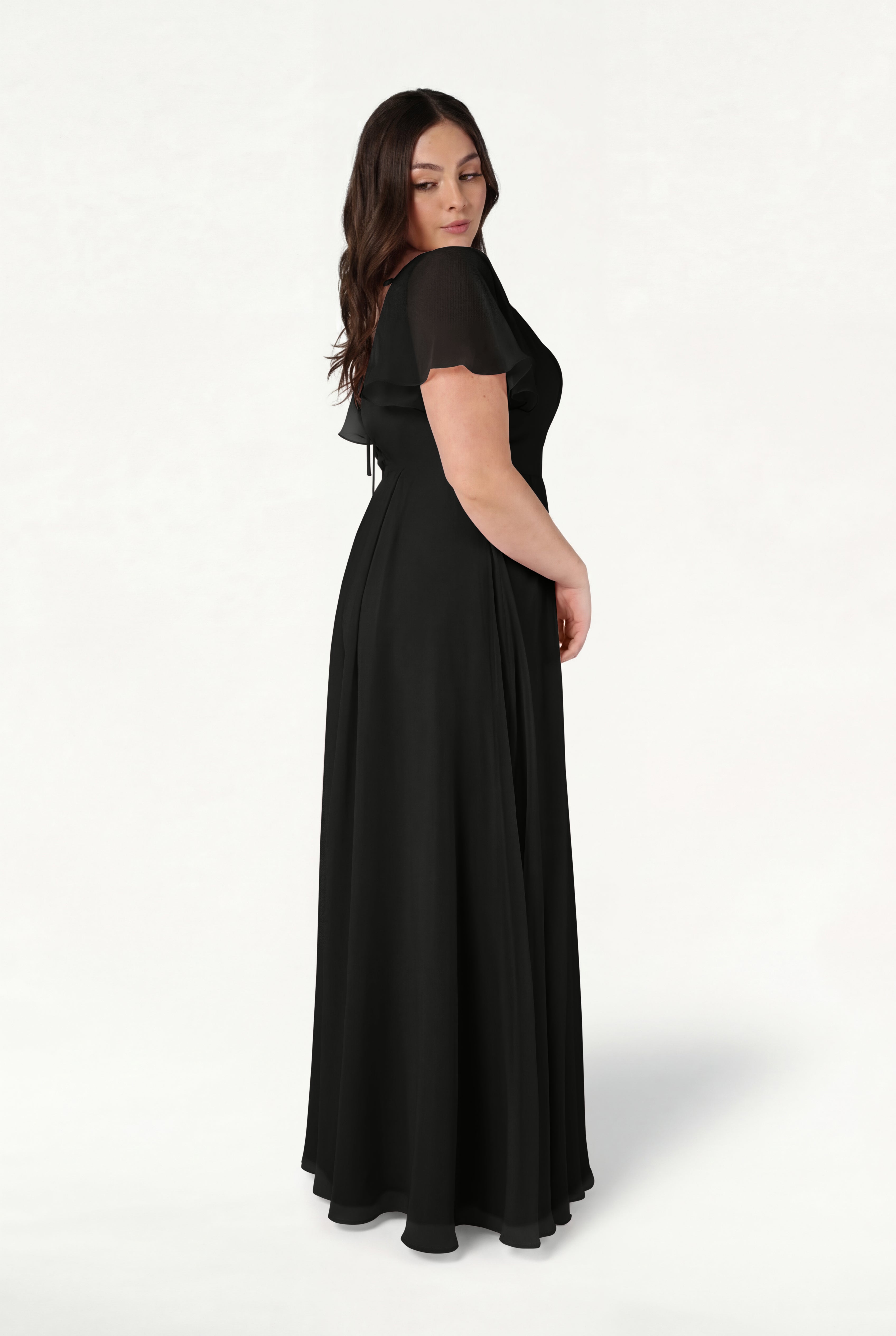 A woman with long brown hair stands sideways against a plain white background, modeling the TH&TH Phoebe Bridesmaid Dress in black chiffon—a floor-length gown with short, sheer sleeves.