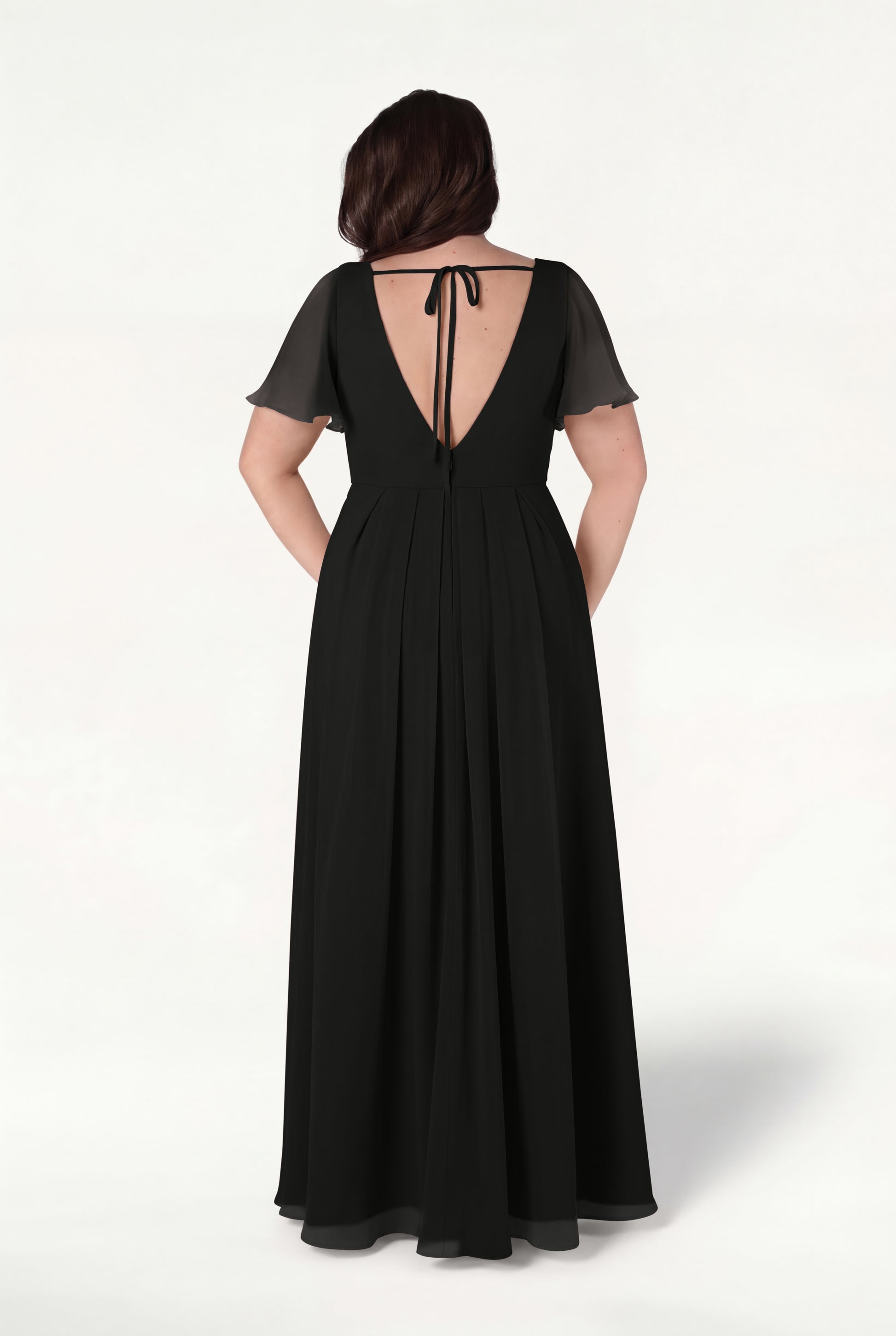 A woman stands facing away on a light background wearing the Phoebe Bridesmaid Dress in Black Chiffon by TH&TH, featuring a floor-length silhouette, short sheer sleeves, and a deep V-back tied at the neck with a ribbon.