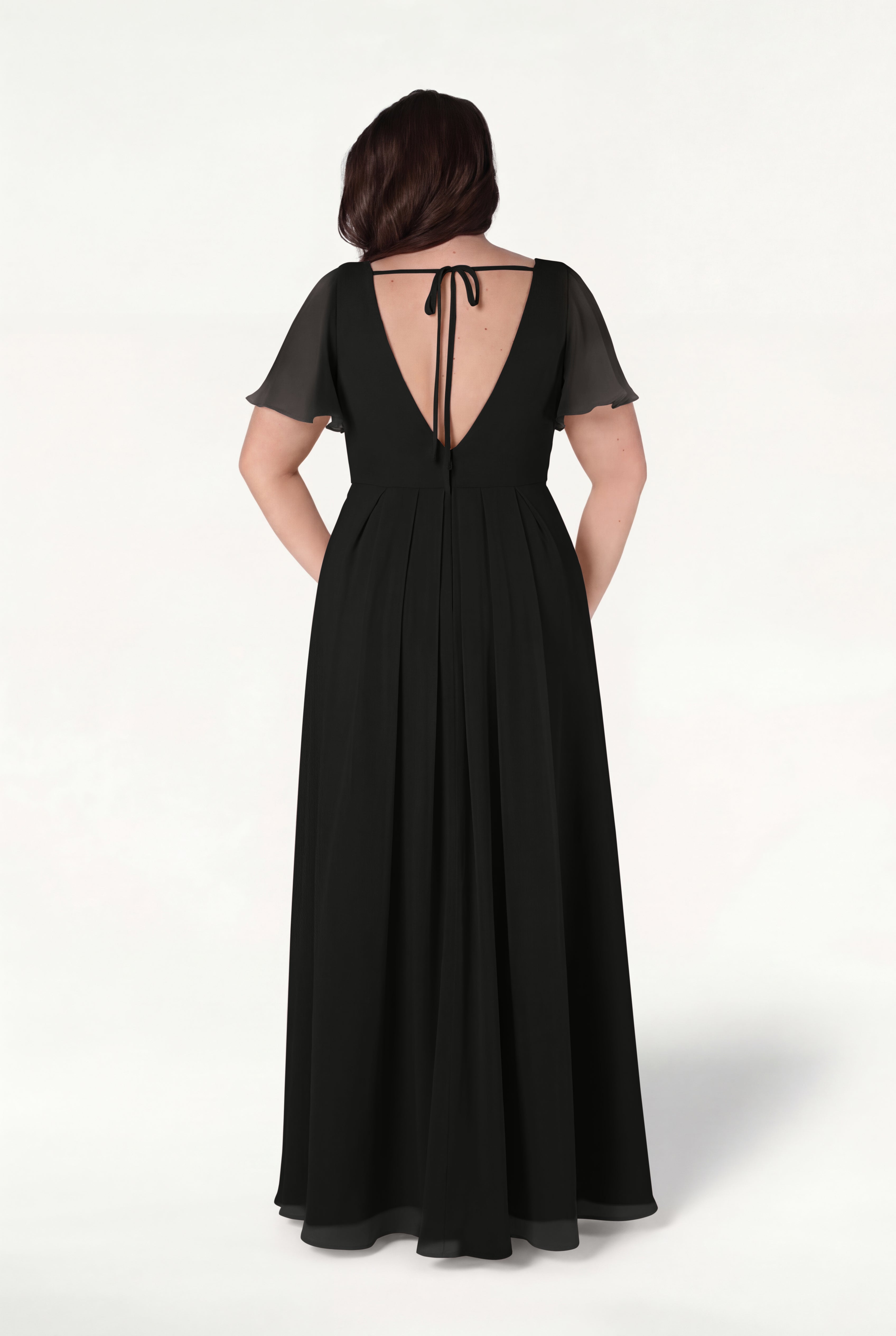 A woman stands facing away on a light background wearing the Phoebe Bridesmaid Dress in Black Chiffon by TH&TH, featuring a floor-length silhouette, short sheer sleeves, and a deep V-back tied at the neck with a ribbon.