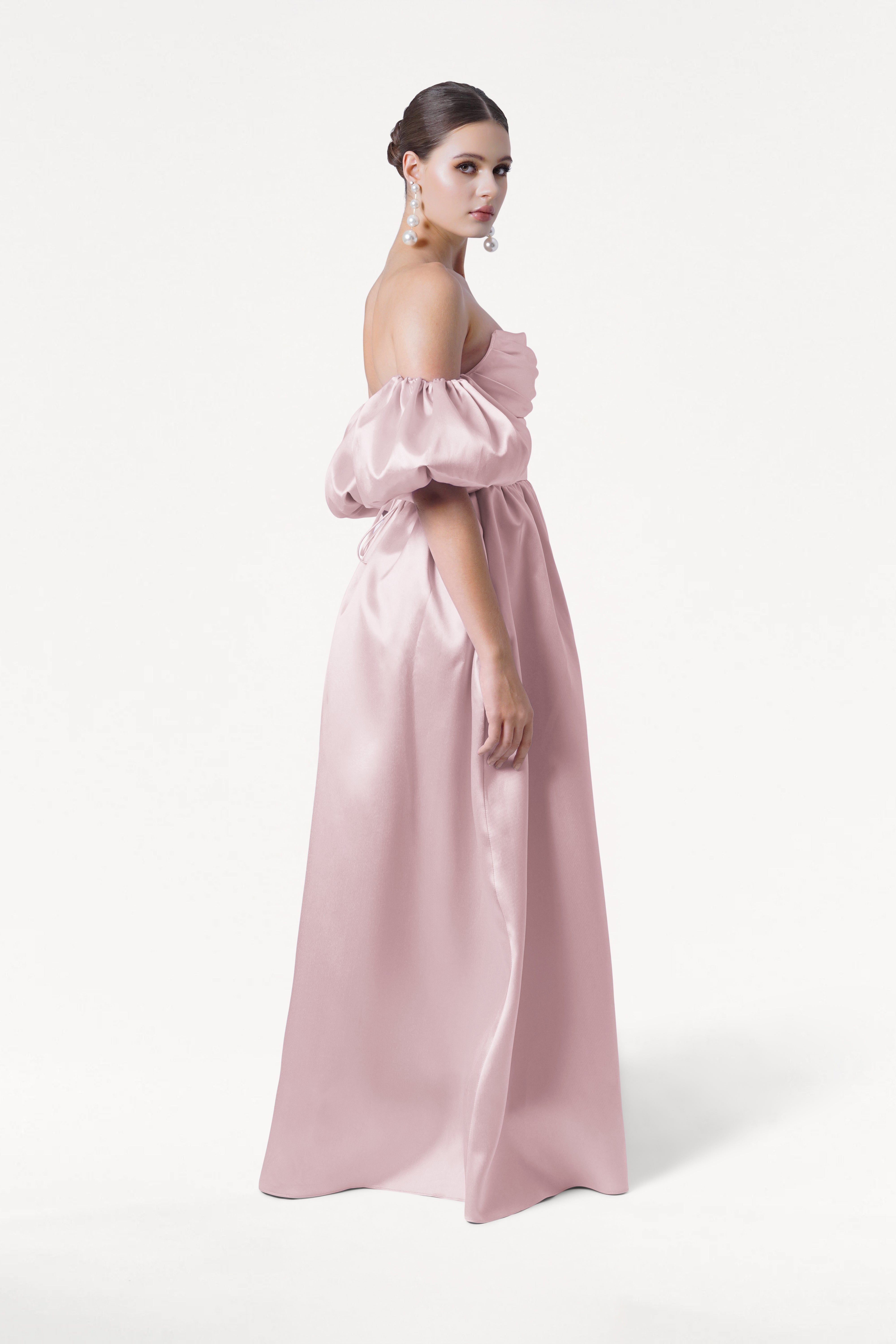 A woman stands sideways in the TH&TH Blair Bridesmaid Dress, a long off-the-shoulder blush pink satin twill gown with detachable puffed sleeves. She wears pearl earrings and has her hair in a low bun against a plain white background.