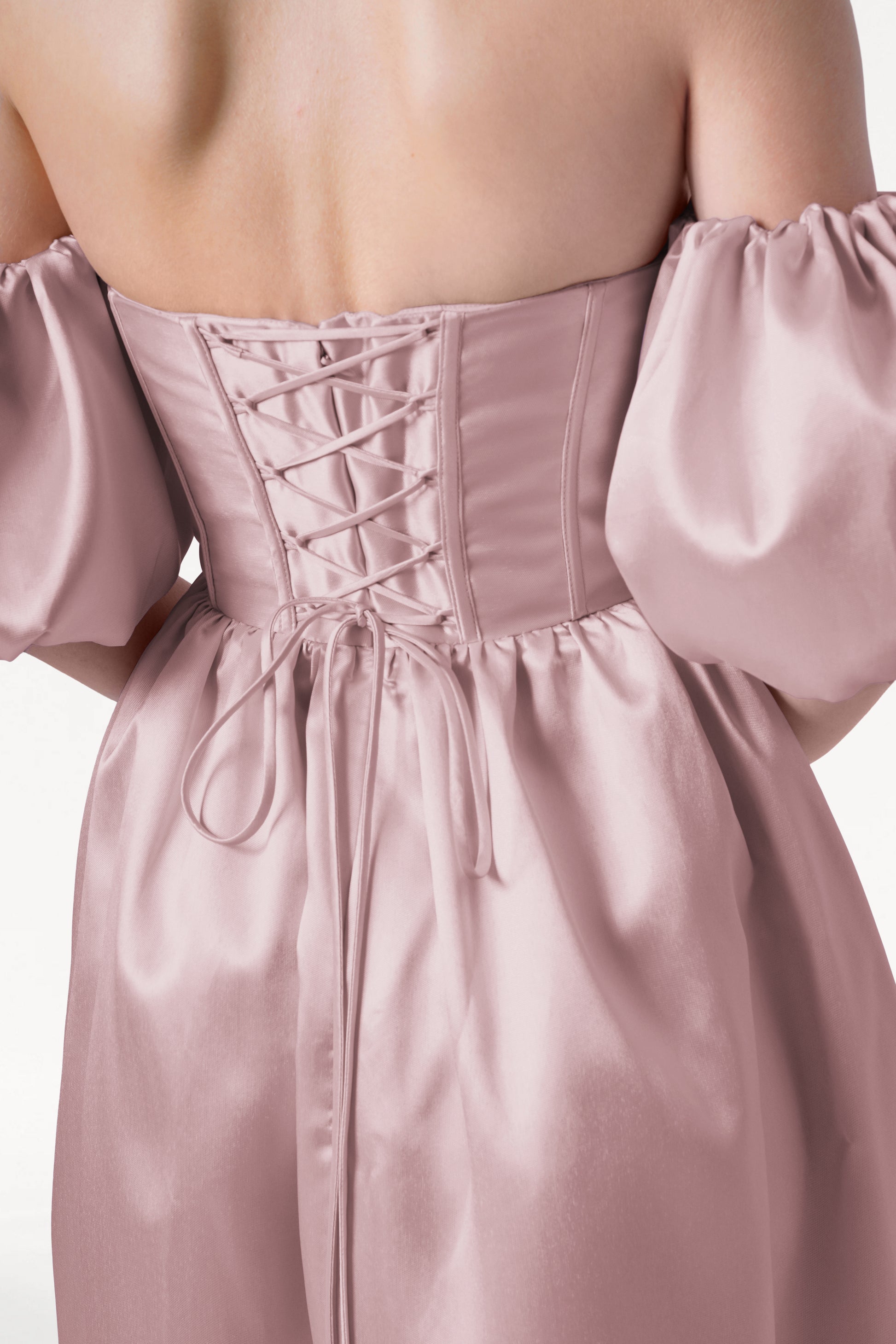 A woman wearing the Blair Bridesmaid Dress in Smoked Blush Satin Twill by TH&TH is shown from the back. The off-shoulder gown features detachable puff sleeves and a corset-style lace-up back, adding romantic, wedding-inspired detail.