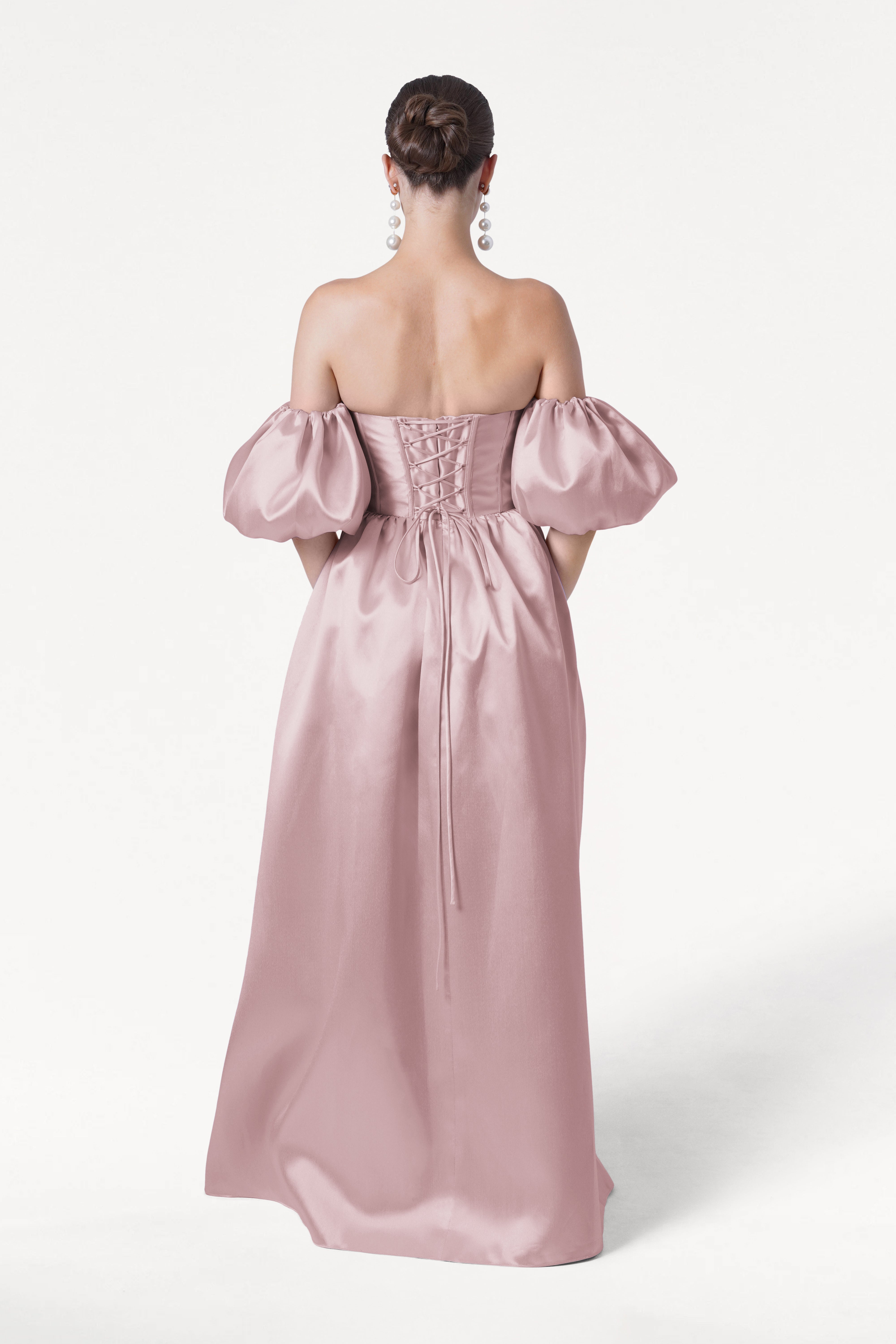 A woman stands facing away in the Blair Bridesmaid Dress by TH&TH, featuring smoked blush satin twill. Her hair is in a low bun, and she wears large dangling earrings, all set against a plain white background.