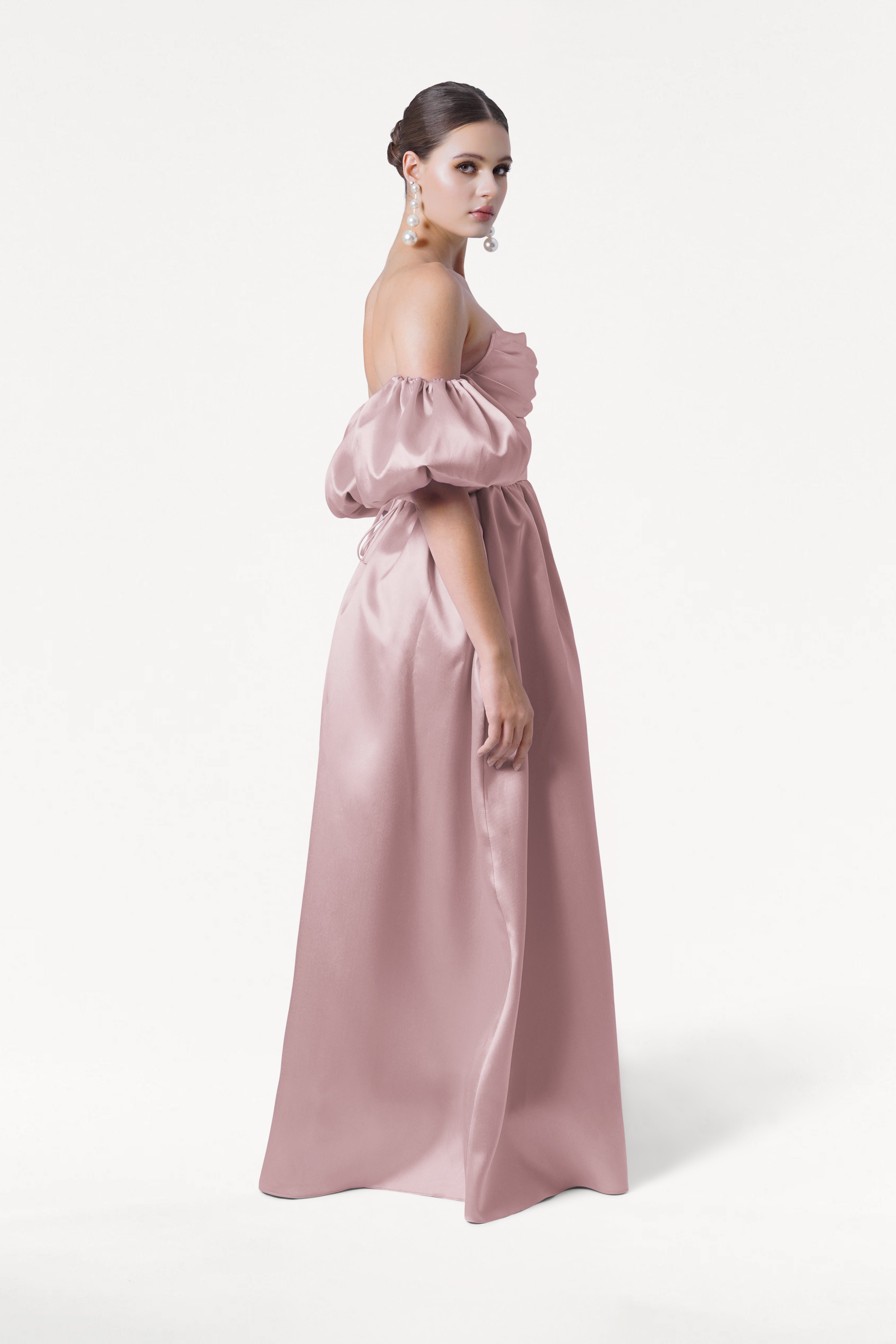 A woman wears the TH&TH Blair Bridesmaid Dress in Smoked Blush Satin Twill with detachable puff sleeves, posing against a white background. Her hair is in a low bun, and she accessorizes with large pearl earrings.