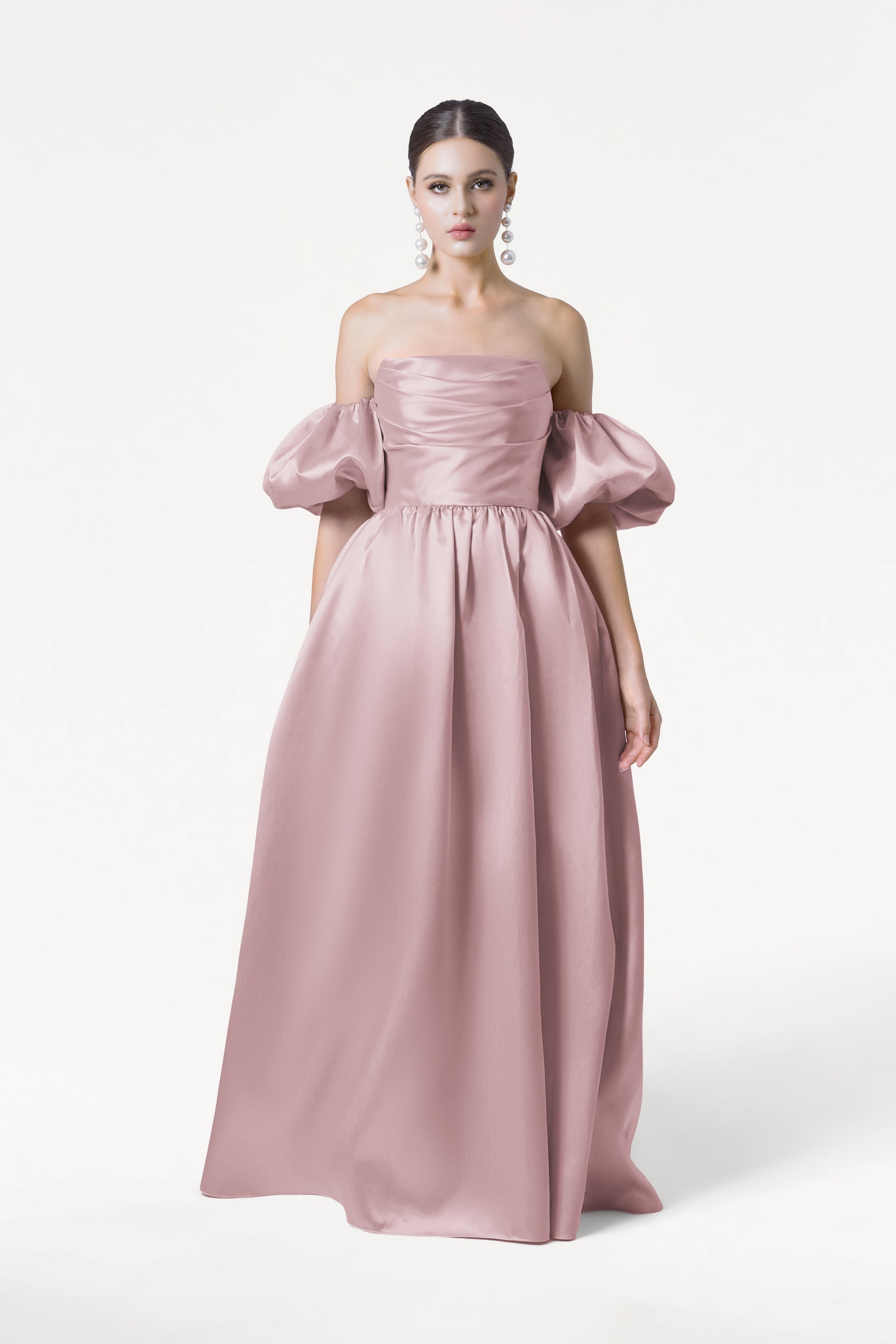 A woman with dark hair pulled back and large dangling earrings poses against a white background, wearing the TH&TH Blair Bridesmaid Dress in Smoked Blush Satin Twill—an off-the-shoulder pink gown with detachable puff sleeves.