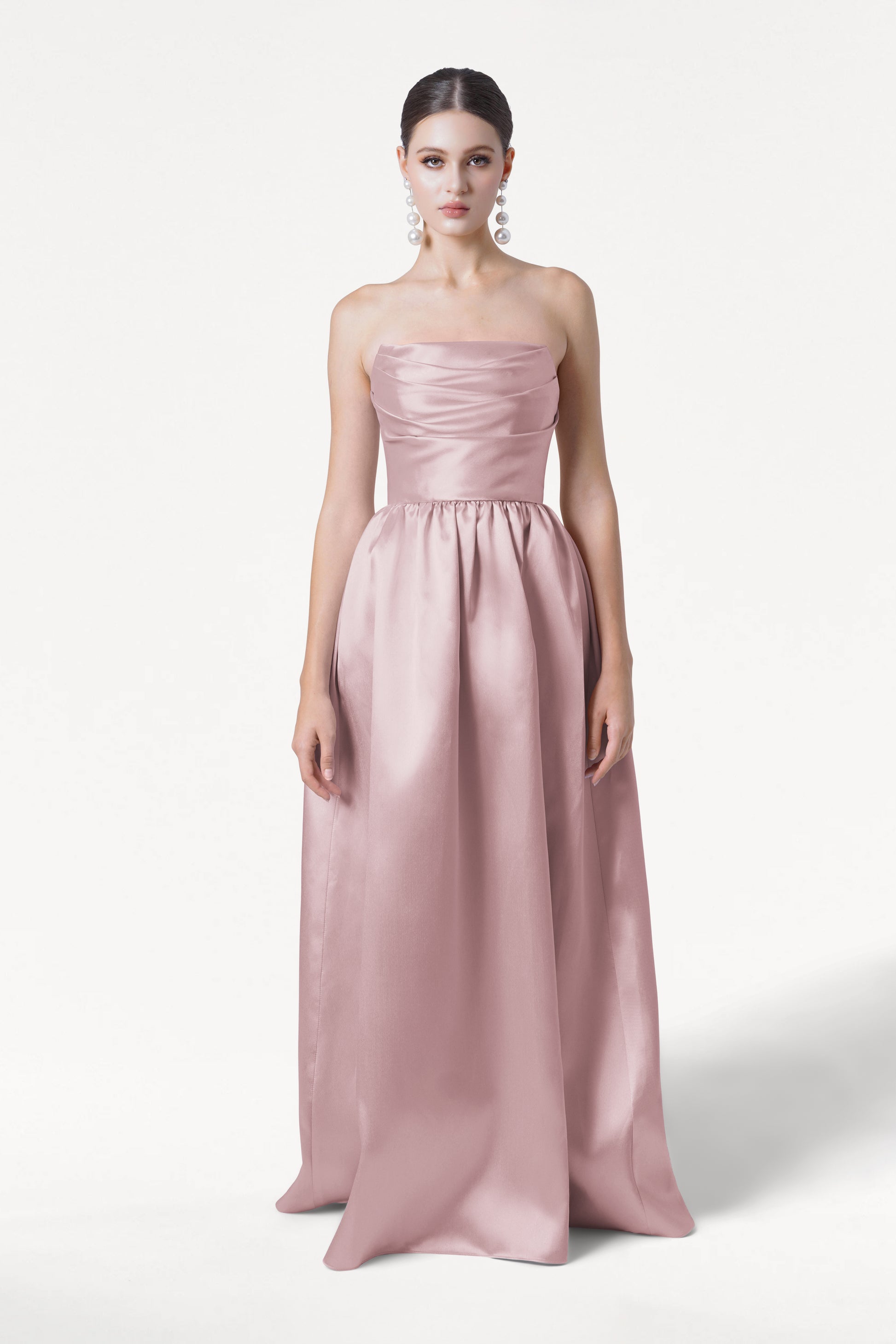 A woman stands against a white background wearing the TH&TH Blair Bridesmaid Dress in Smoked Blush Satin Twill, a strapless, floor-length gown with a fitted bodice and gathered skirt. Her hair is pulled back and she wears large statement earrings.
