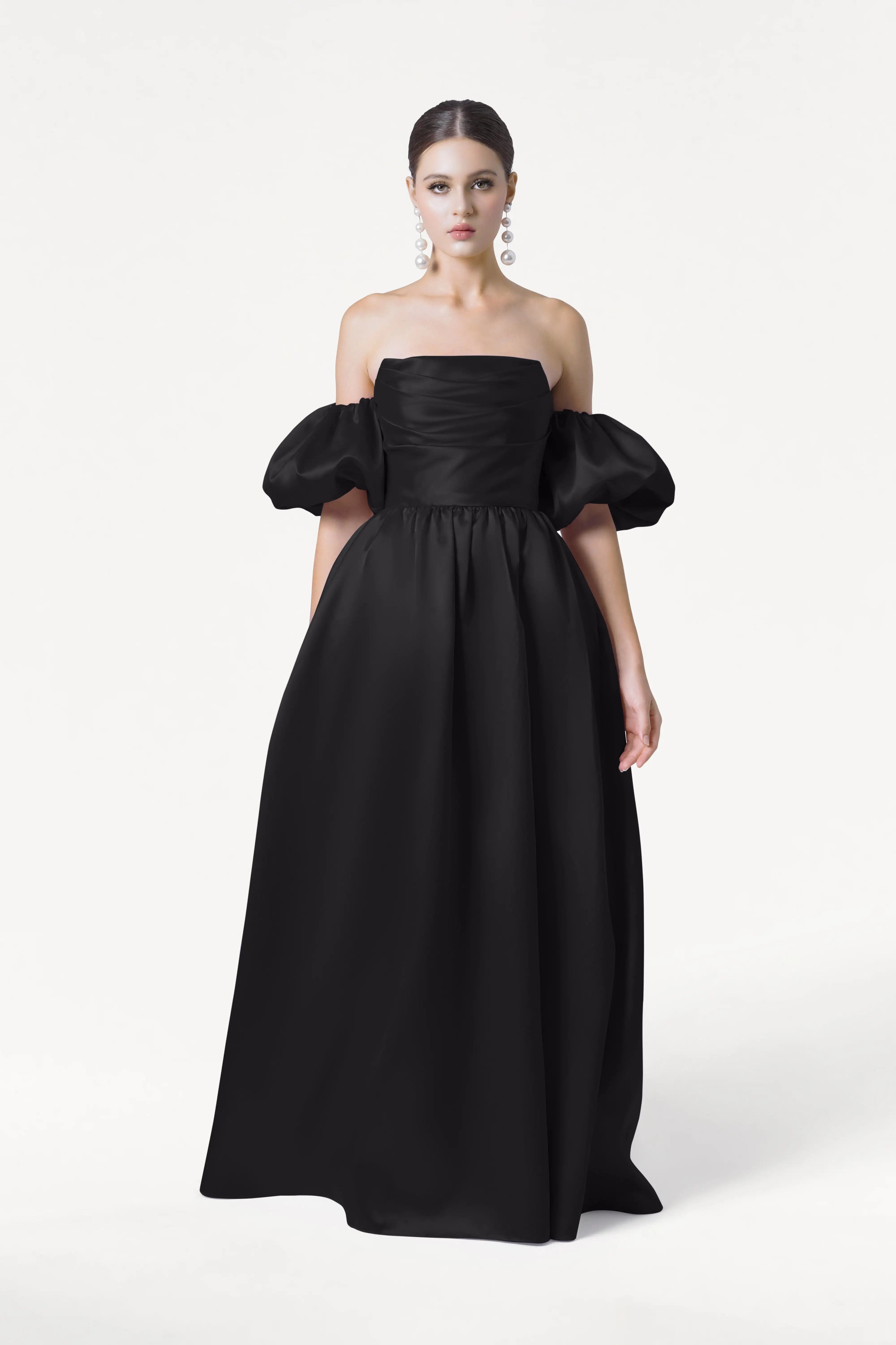 A woman poses against a white background wearing the TH&TH Blair Bridesmaid Dress in Black Satin Twill, a long off-shoulder gown with puffed short sleeves. Her dark hair is pulled back and she accessorizes with large, dangling earrings.