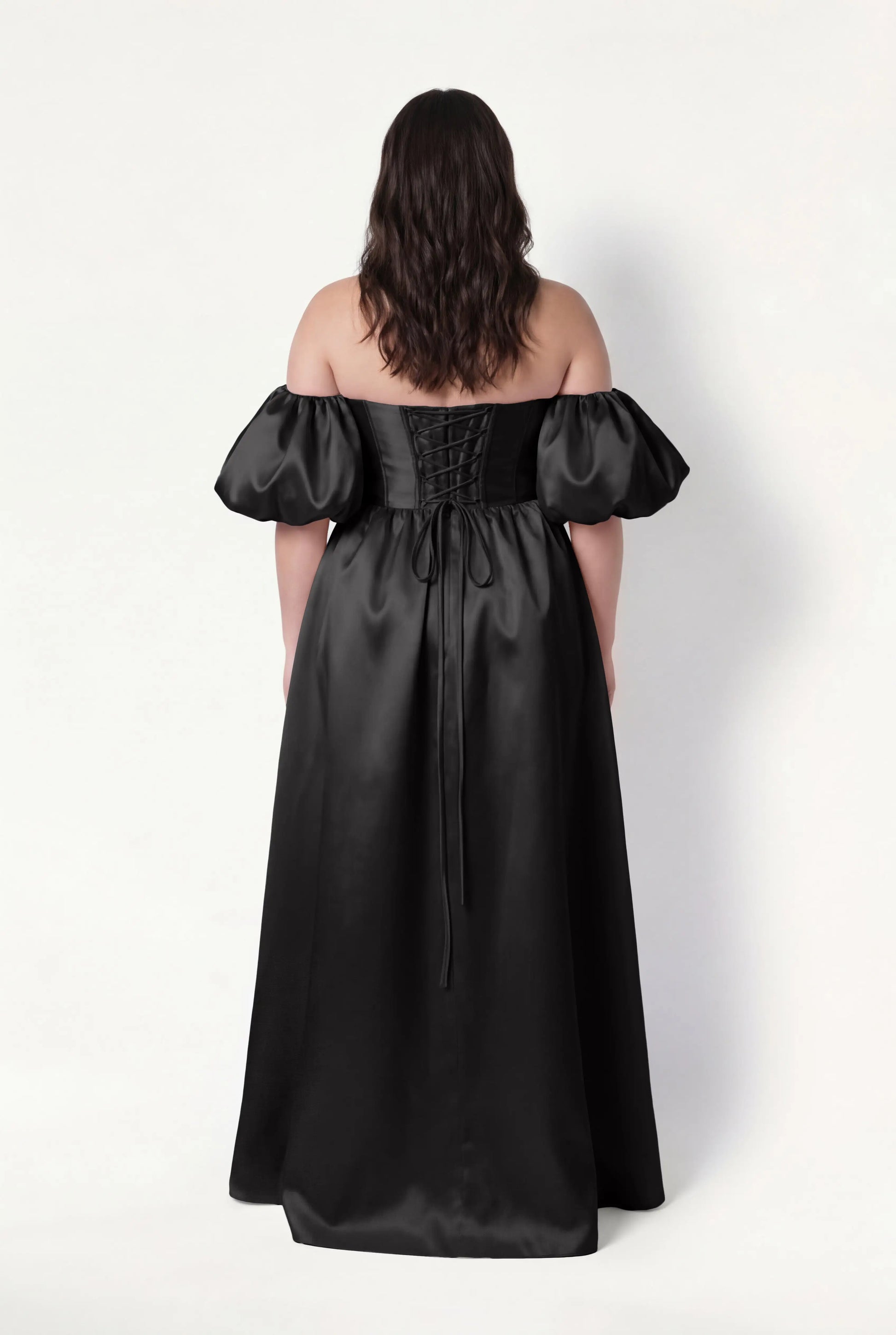 A woman with wavy brown hair is shown from behind wearing the TH&TH Blair Bridesmaid Dress in Black Satin Twill, featuring an off-the-shoulder corset bodice, puffed detachable sleeves, lace-up back, and a floor-length skirt. Plain white background.