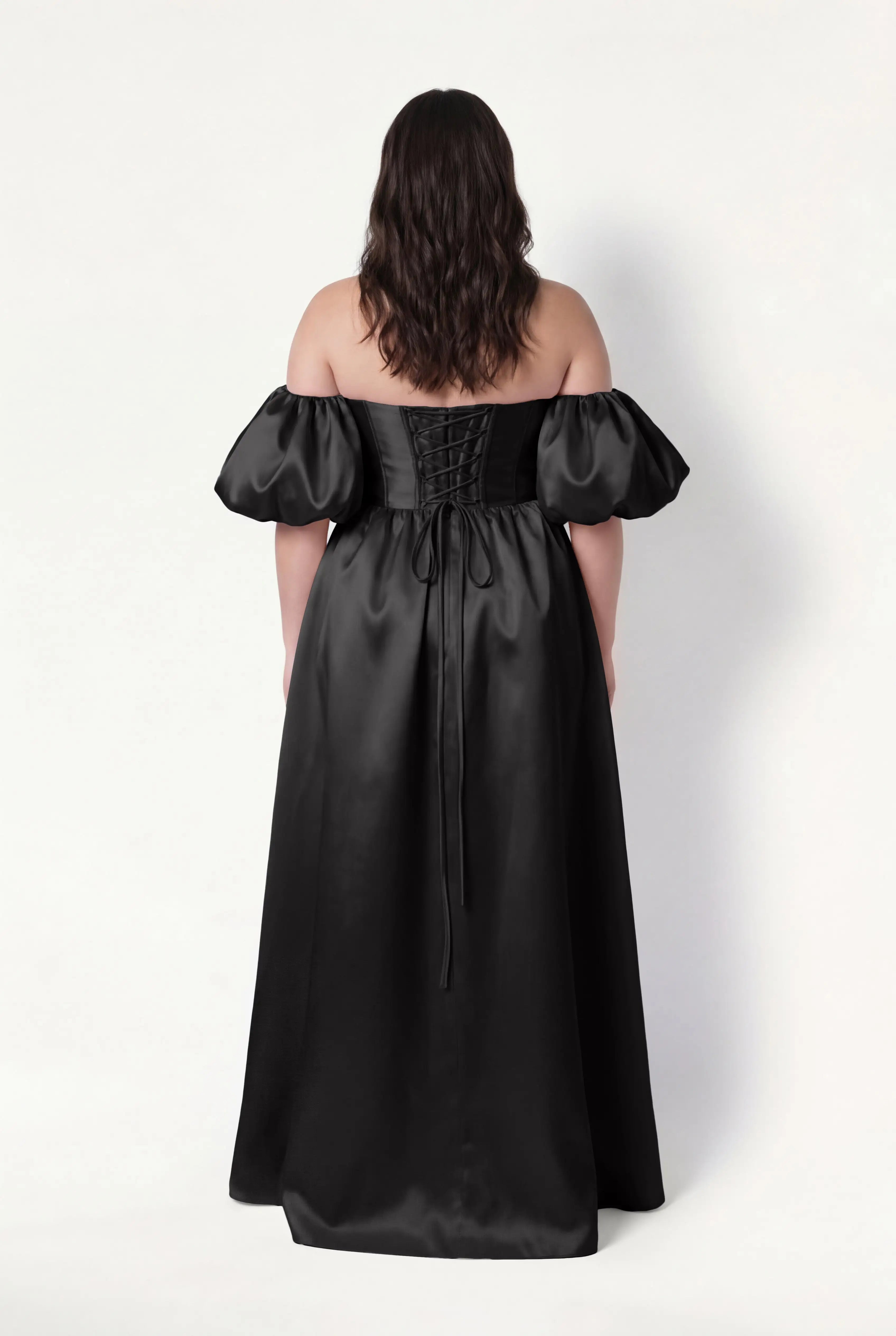 A woman with wavy brown hair is shown from behind wearing the TH&TH Blair Bridesmaid Dress in Black Satin Twill, featuring an off-the-shoulder corset bodice, puffed detachable sleeves, lace-up back, and a floor-length skirt. Plain white background.