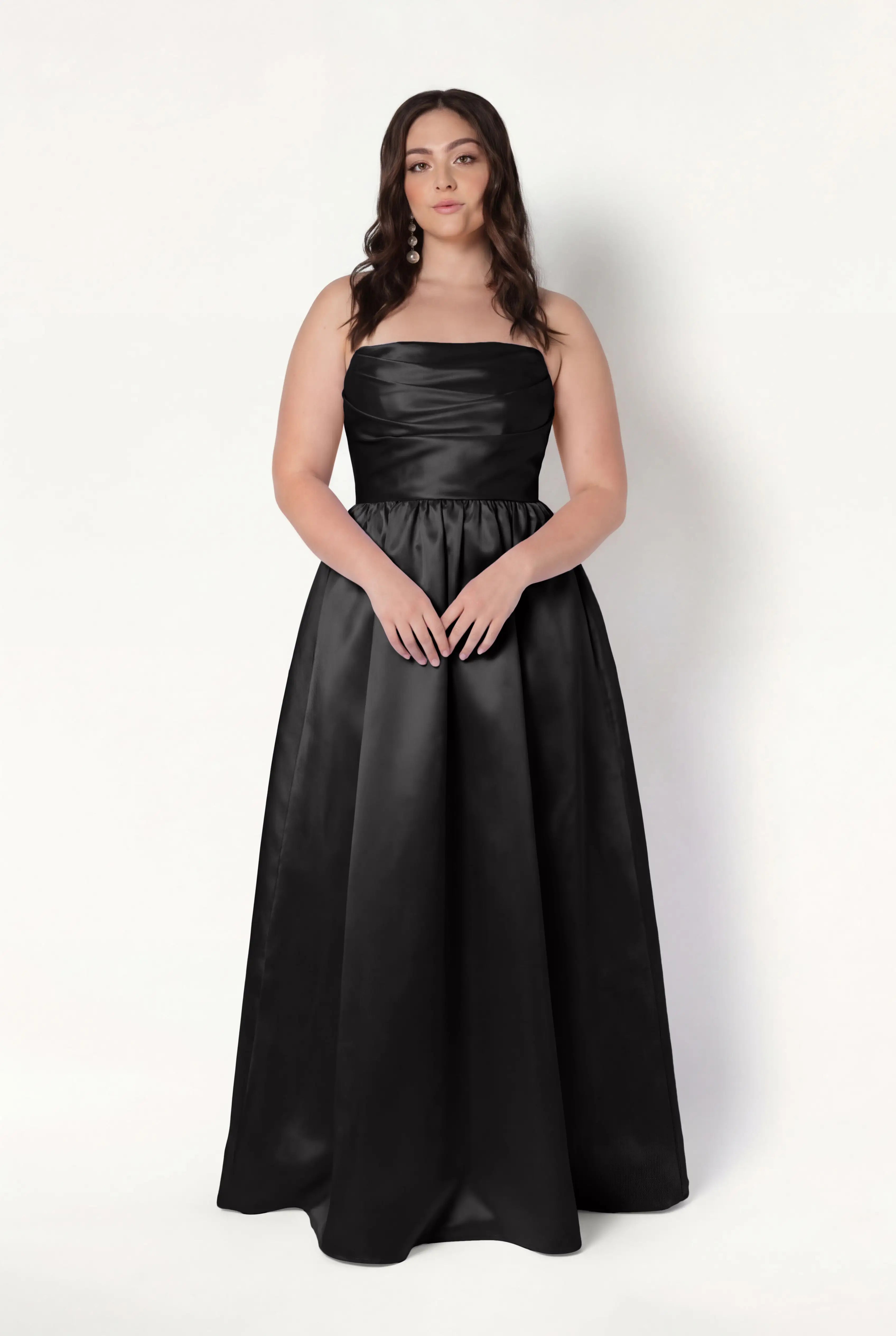 A woman with long brown hair models the TH&TH Blair Bridesmaid Dress in Black Satin Twill, a strapless, floor-length gown, against a plain white background.