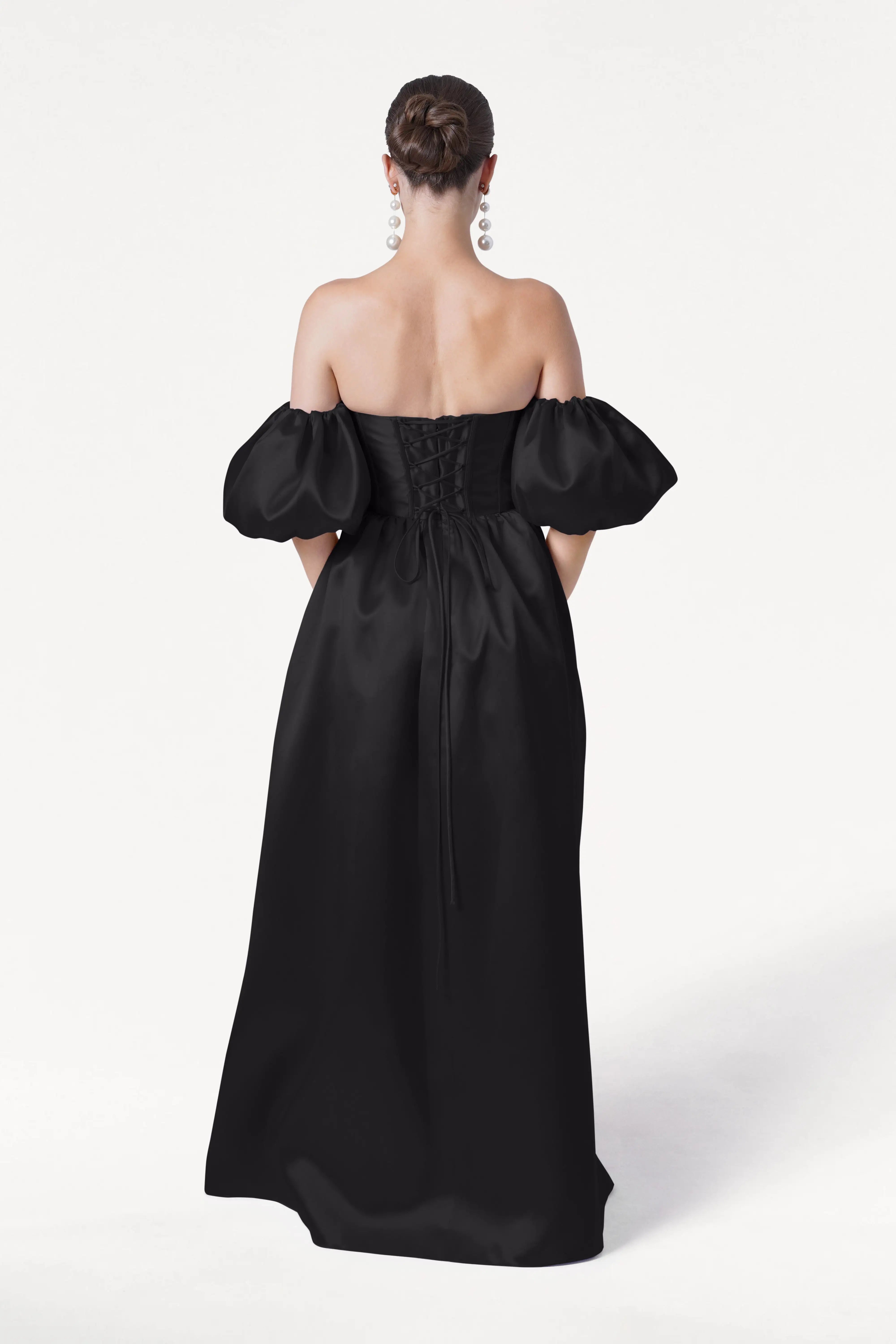 A woman with her hair in a neat bun and large dangling earrings wears the TH&TH Blair Bridesmaid Dress in Black Satin Twill, an off-the-shoulder corset gown with puffed sleeves and lace-up back, set against a plain white background.