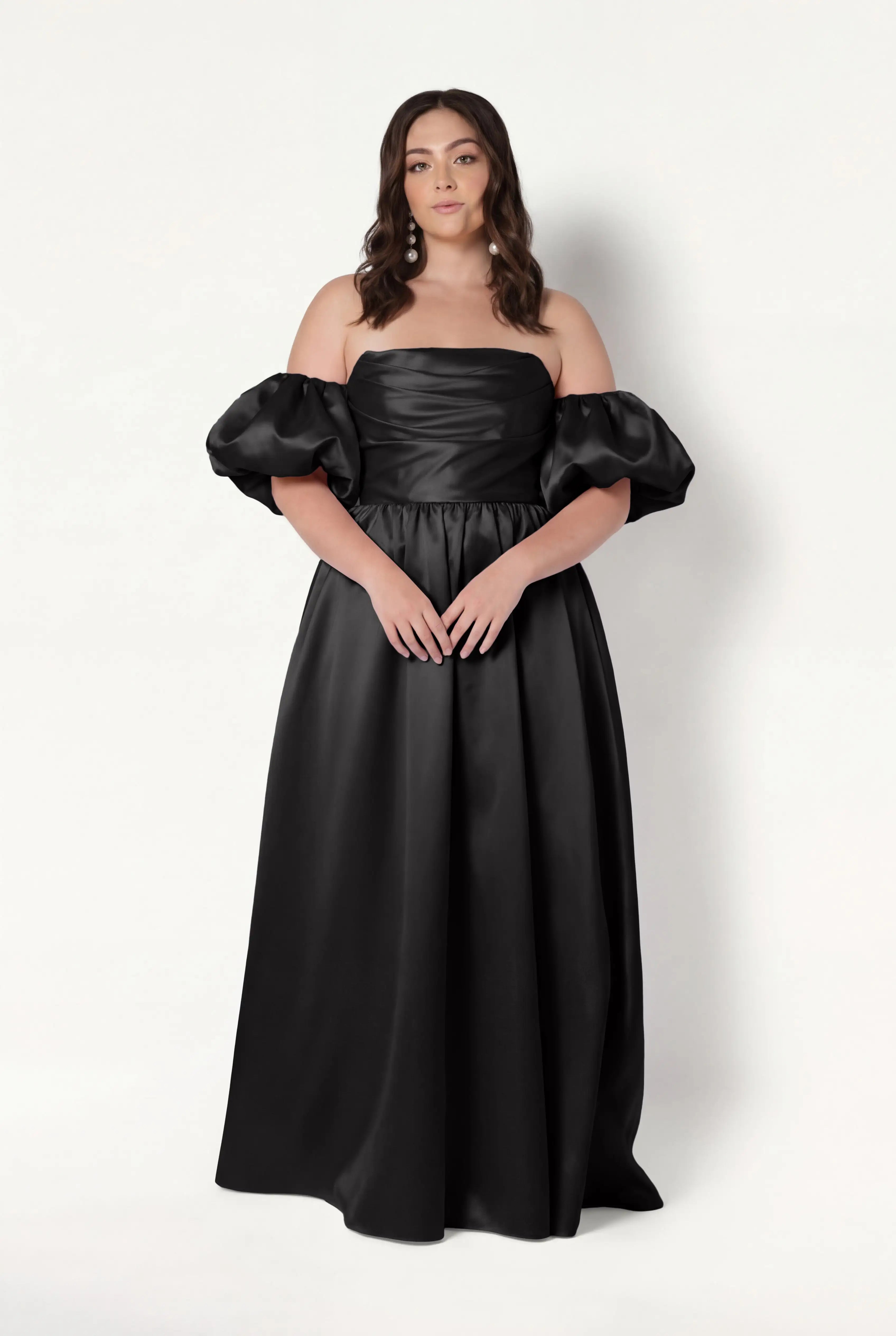 A woman with wavy brown hair and a neutral expression stands against a white background wearing the TH&TH Blair Bridesmaid Dress in Black Satin Twill, featuring an off-the-shoulder neckline, puffed short sleeves, and a fitted corset bodice.