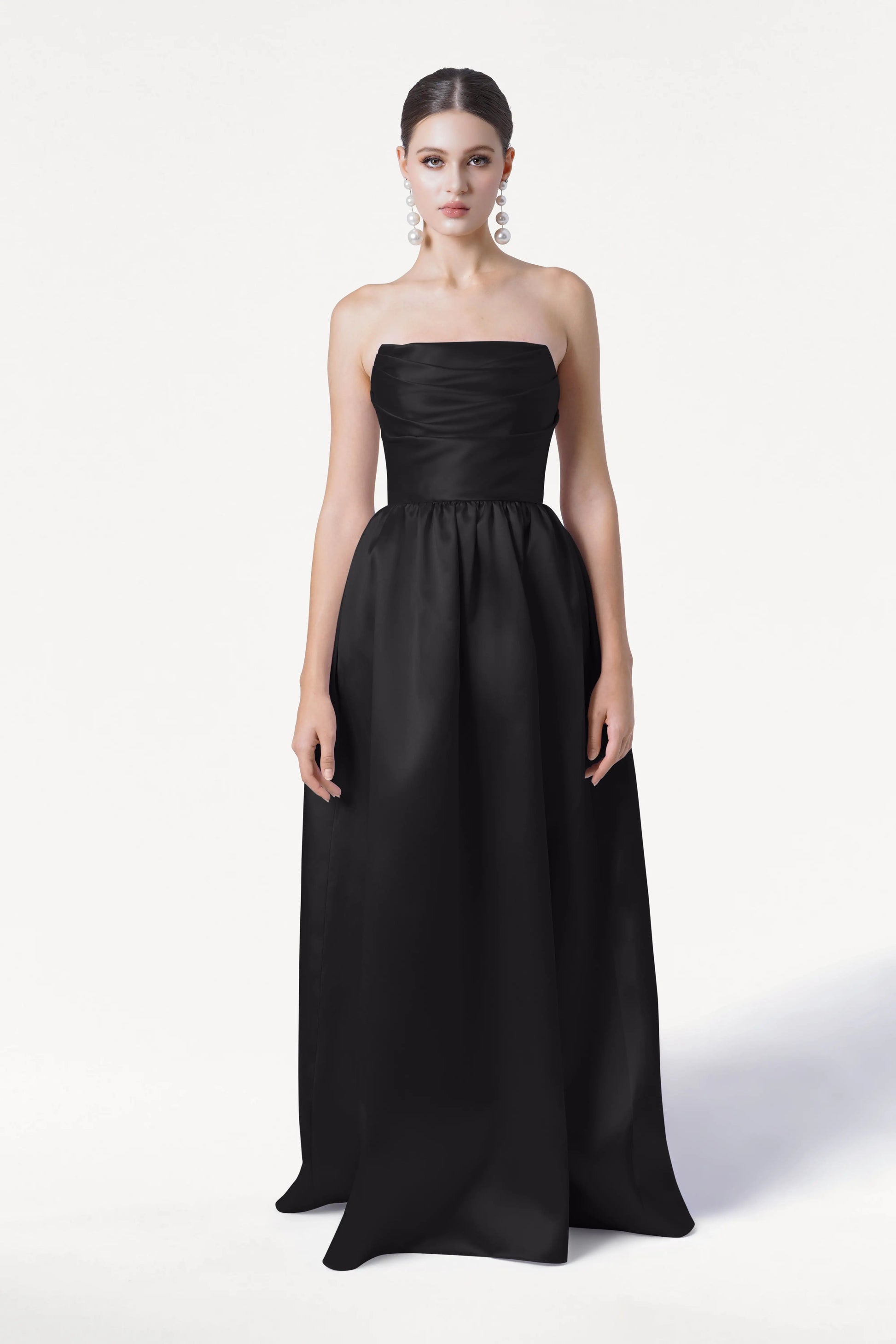A woman faces forward, wearing the TH&TH Blair Bridesmaid Dress in Black Satin Twill. The strapless, floor-length gown features a fitted bodice and full skirt. Her hair is pulled back, she wears large earrings, and the background is plain white.
