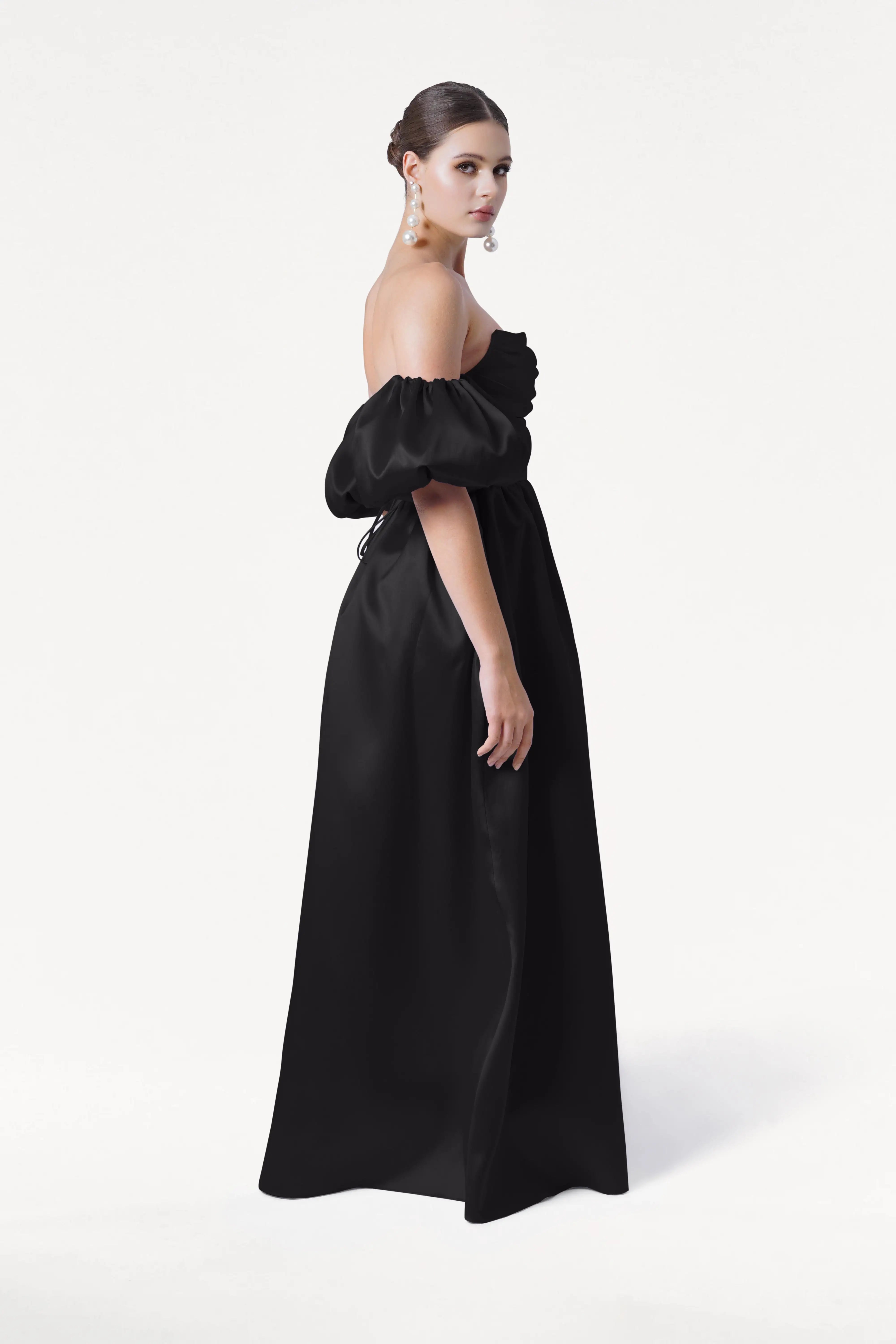 A woman stands sideways against a white background, wearing the Blair Bridesmaid Dress in Black Satin Twill by TH&TH with detachable puffed sleeves. Her hair is in a neat bun and she wears large, dangling earrings.