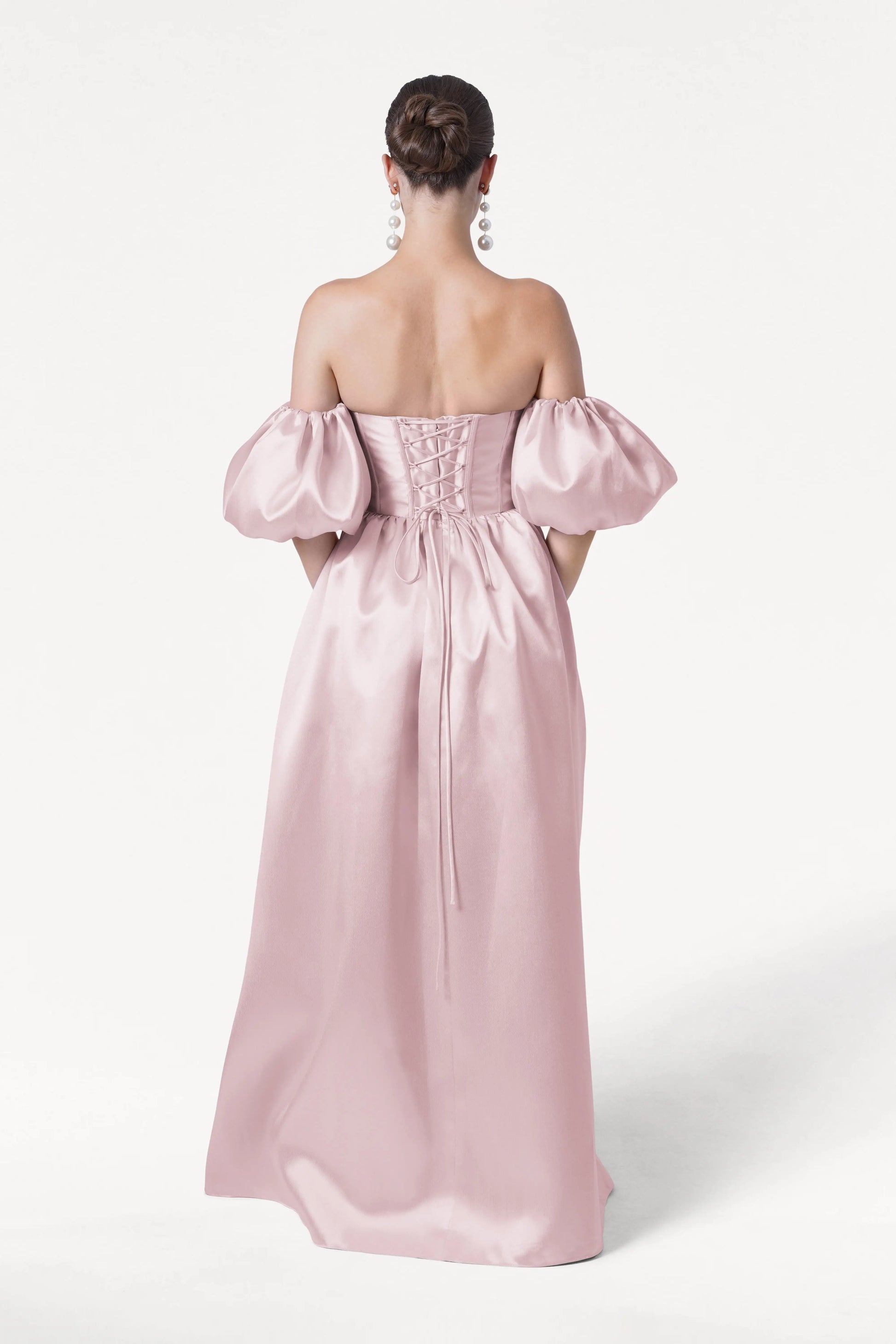 A woman with an elegant bun and long earrings faces away, wearing the TH&TH Blair Bridesmaid Dress in blush pink satin twill, featuring an off-the-shoulder design, puffed detachable sleeves, and a lace-up back against a plain white background.
