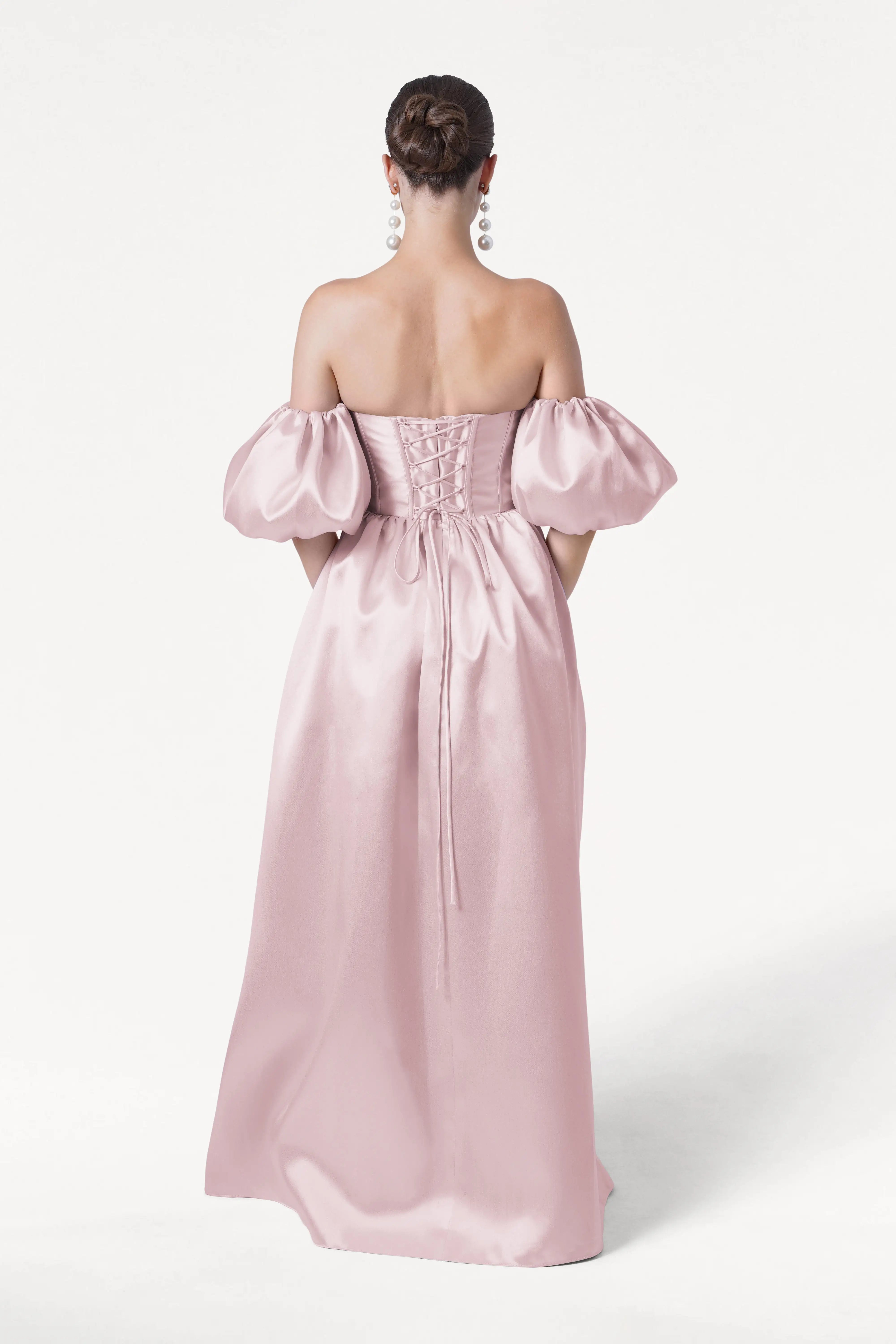 A woman with an elegant bun and long earrings faces away, wearing the TH&TH Blair Bridesmaid Dress in blush pink satin twill, featuring an off-the-shoulder design, puffed detachable sleeves, and a lace-up back against a plain white background.