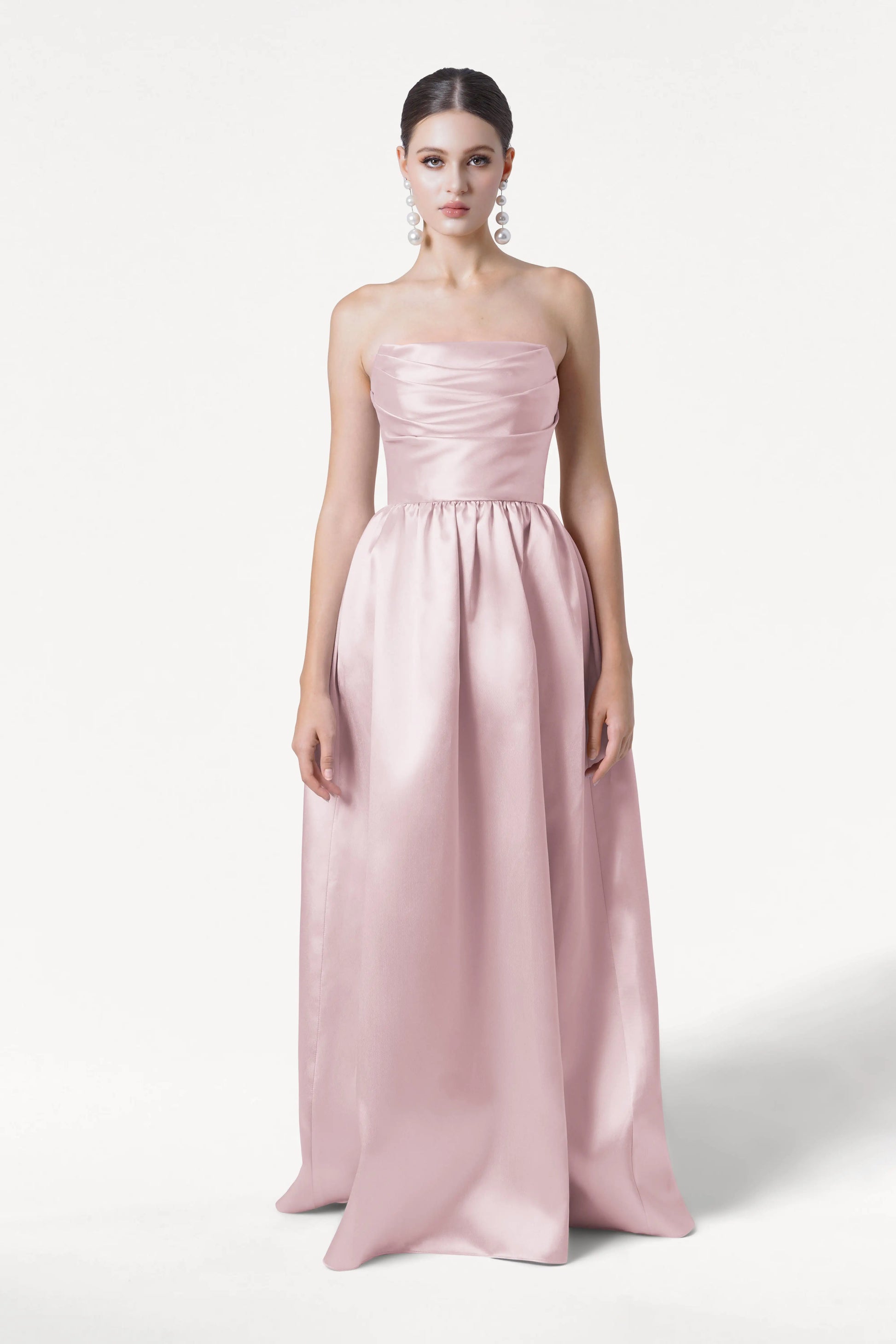 A woman in the Blair Bridesmaid Dress by TH&TH, a strapless, floor-length blush pink satin twill gown, stands facing forward against a white background with her hair pulled back and wearing large pearl earrings.