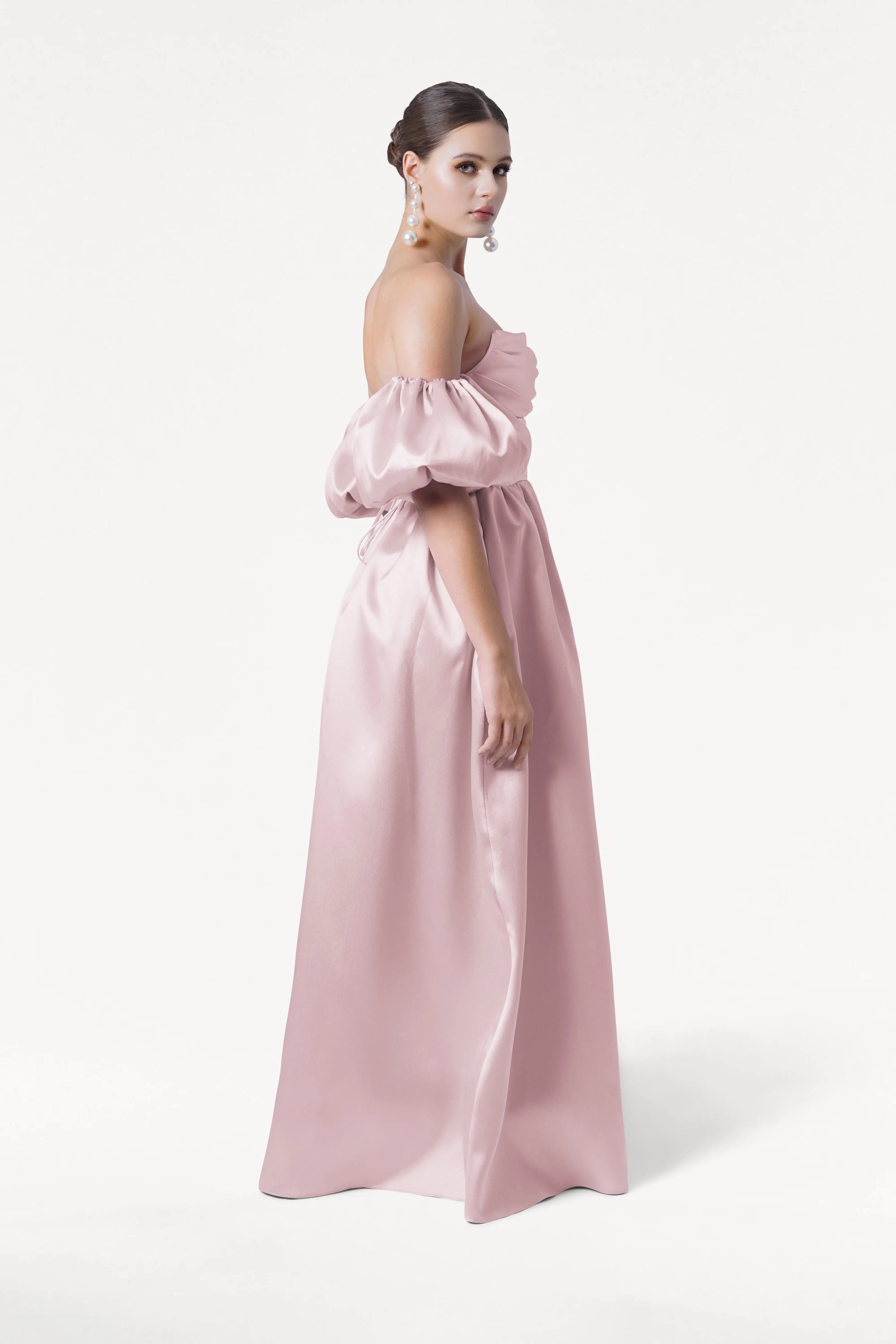 A woman stands sideways in the TH&TH Blair Bridesmaid Dress, a long off-the-shoulder blush pink satin twill gown with detachable puffed sleeves. She wears pearl earrings and has her hair in a low bun against a plain white background.