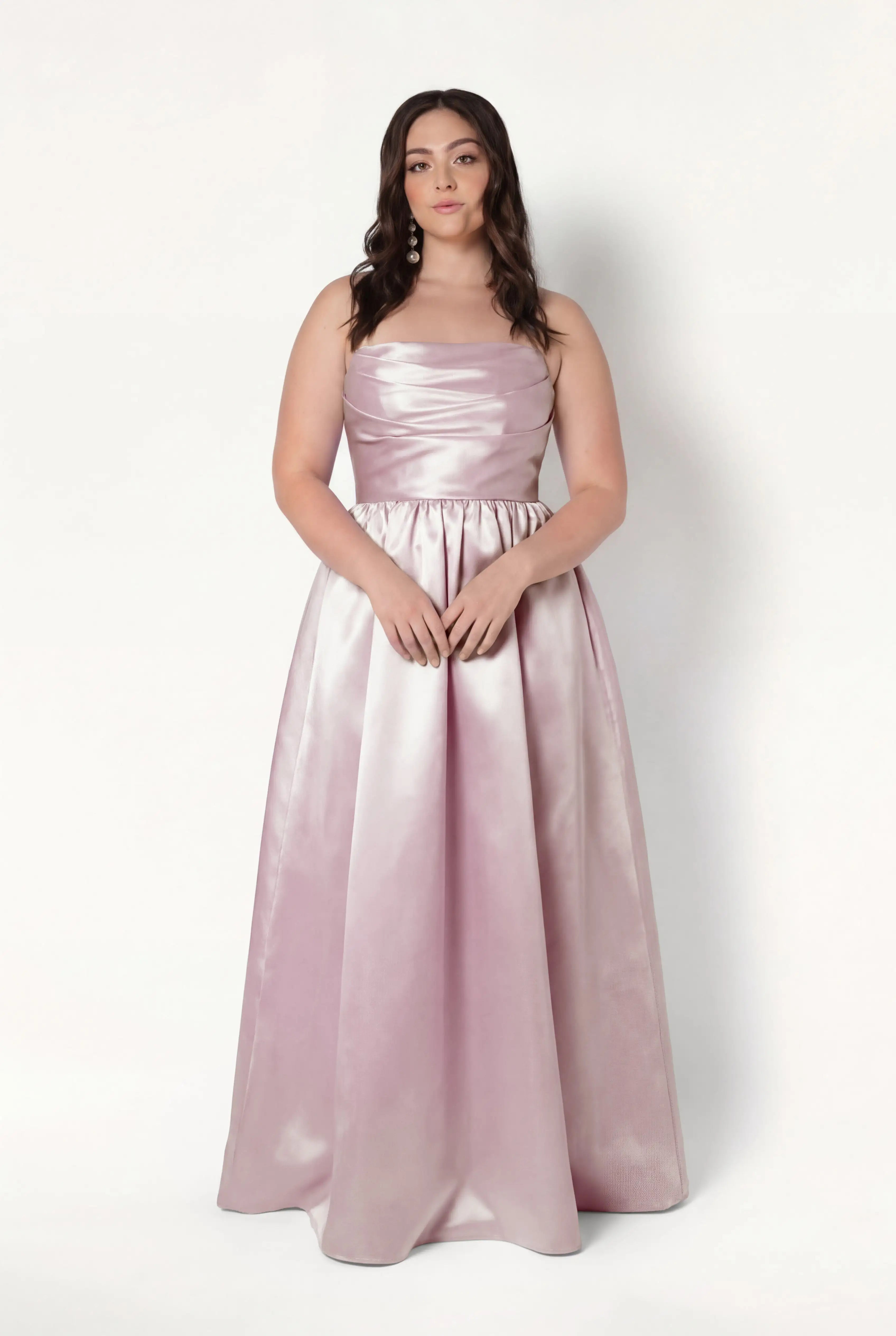 A woman with long, wavy brown hair and a neutral expression wears the TH&TH Blair Bridesmaid Dress in blush pink satin twill, featuring a fitted bodice and full-length skirt, standing against a white background.
