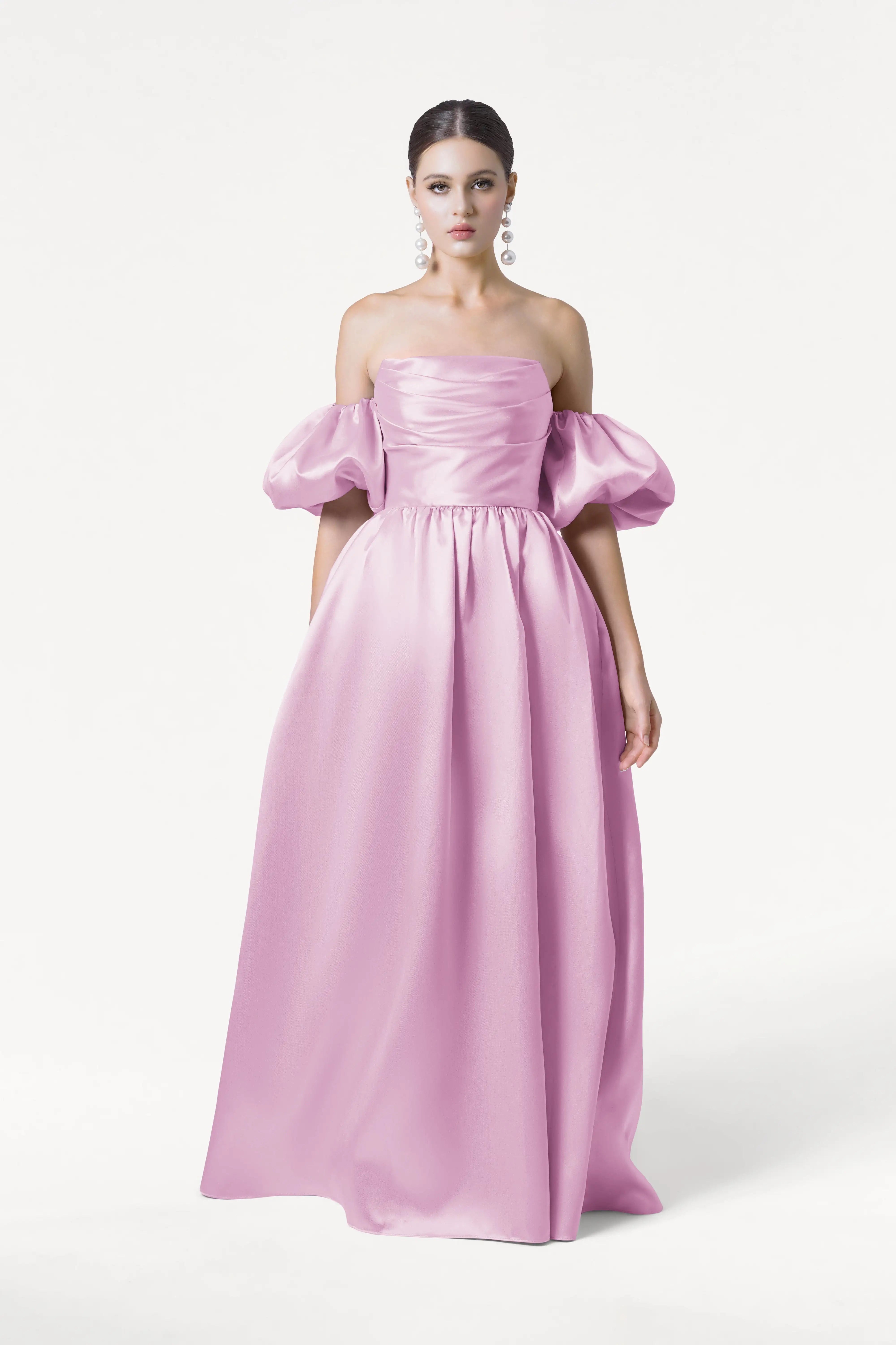 A woman poses against a white backdrop wearing the Blair Bridesmaid Dress in Bubblegum Pink Satin Twill by TH&TH, featuring a floor-length, off-the-shoulder corset design with puff sleeves. Her hair is slicked back and she wears statement earrings.