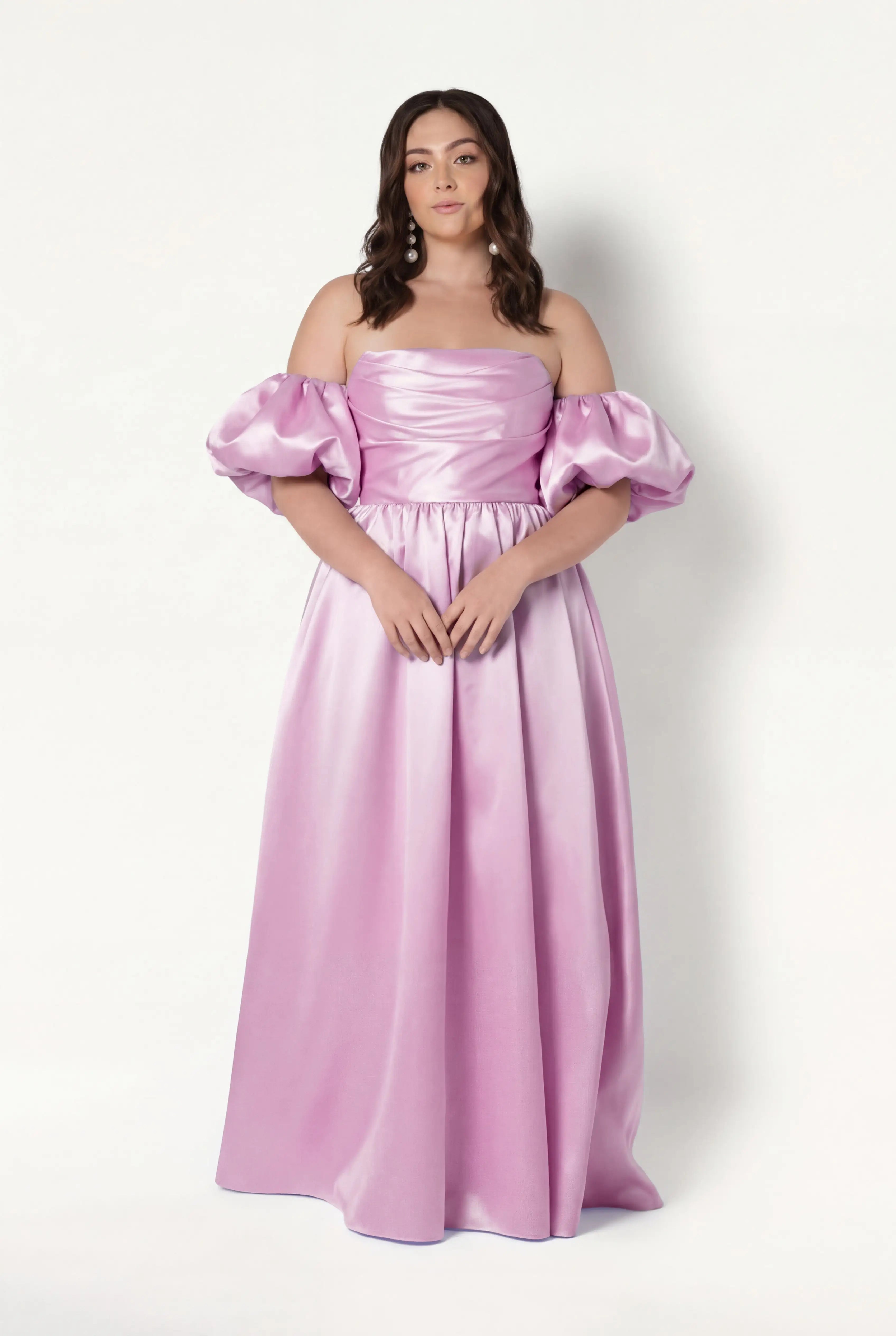 A woman with long, wavy brown hair and a neutral expression poses against a white background in the TH&TH Blair Bridesmaid Dress—an off-the-shoulder, floor-length gown in bubblegum pink satin twill with puffy detachable sleeves.