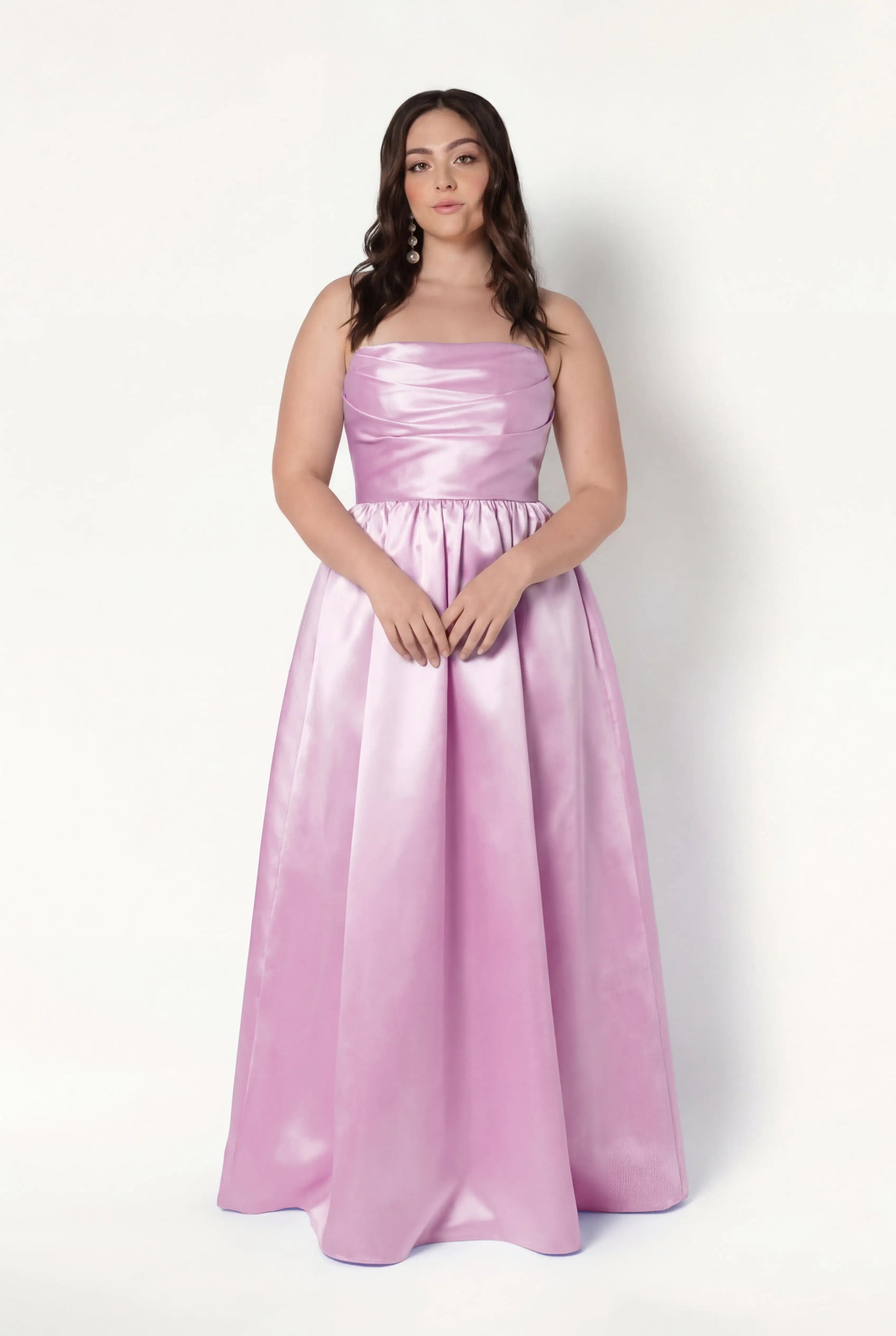 A woman with long brown hair stands against a white background, wearing the TH&TH Blair Bridesmaid Dress in Bubblegum Pink Satin Twill. The shiny, floor-length gown is strapless, and she looks forward with her hands gently clasped in front of her.