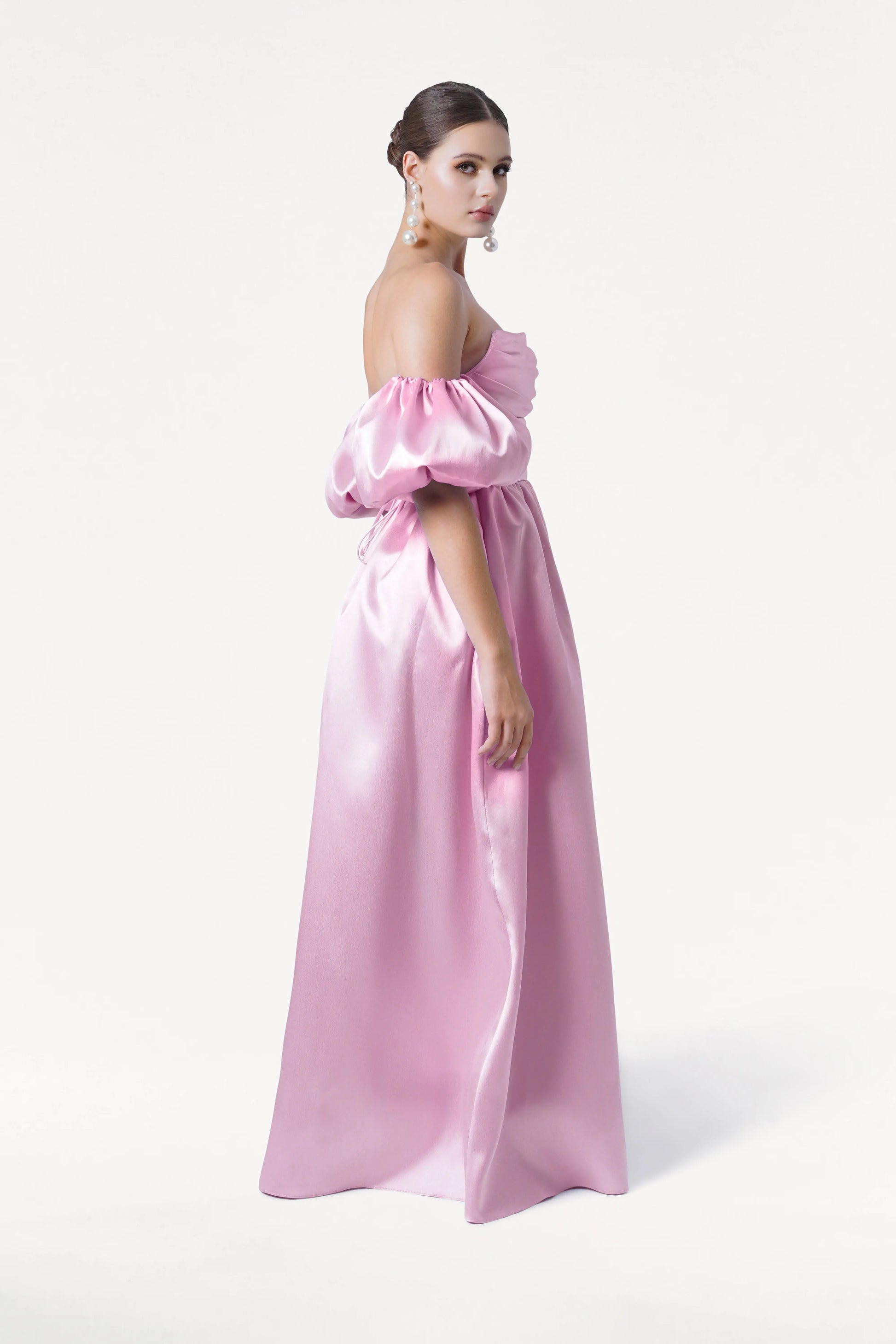 A woman wears the Blair Bridesmaid Dress in Bubblegum Pink Satin Twill by TH&TH, featuring off-shoulder puffed sleeves. She stands on a white background, hair styled up, and accessorized with dangling pearl earrings.