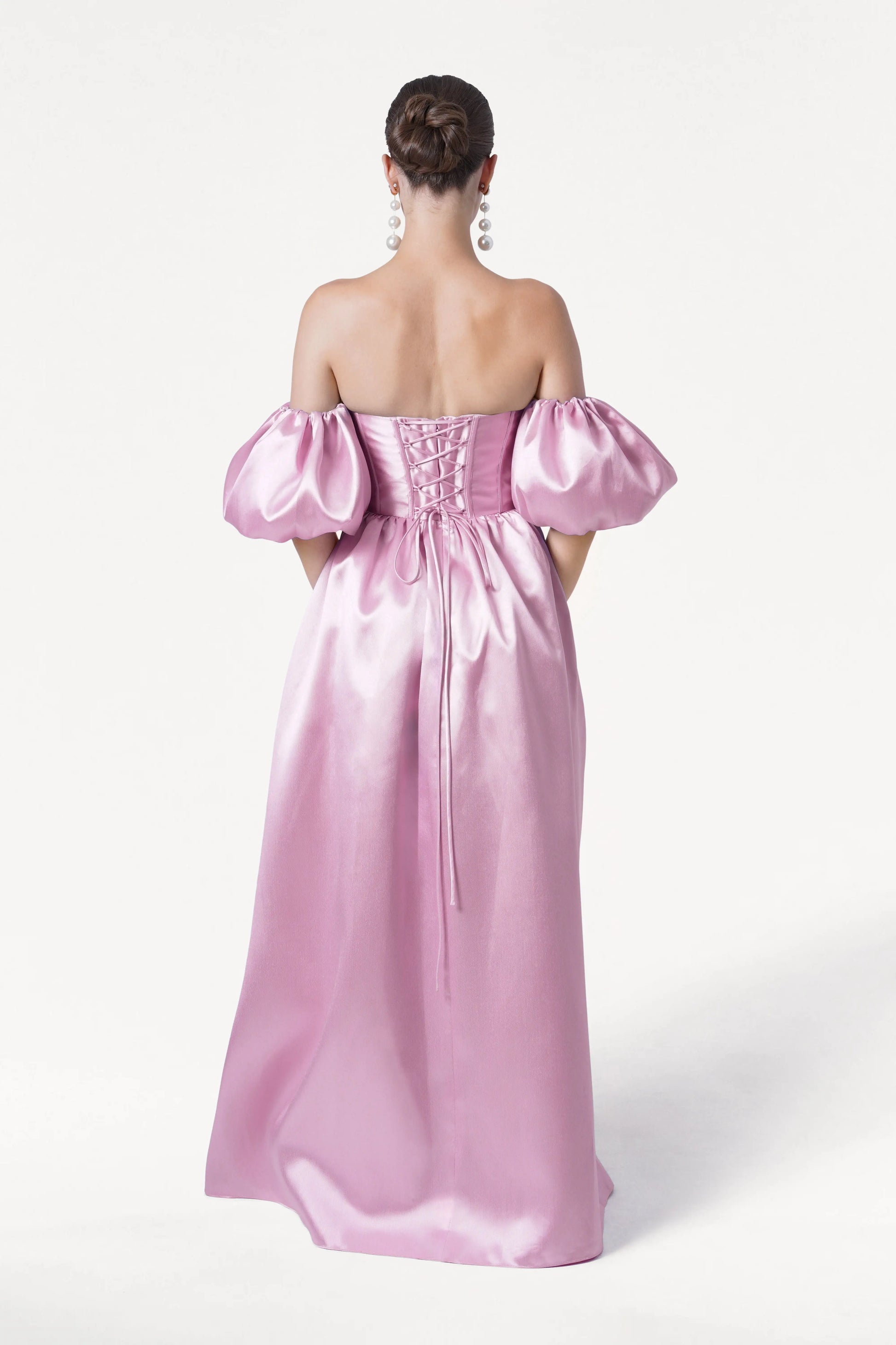 A woman faces away in the TH&TH Blair Bridesmaid Dress, crafted from bubblegum pink satin twill with off-the-shoulder puff sleeves and a lace-up back. Her hair is styled in a neat bun with long pearl earrings, set against a plain white background.
