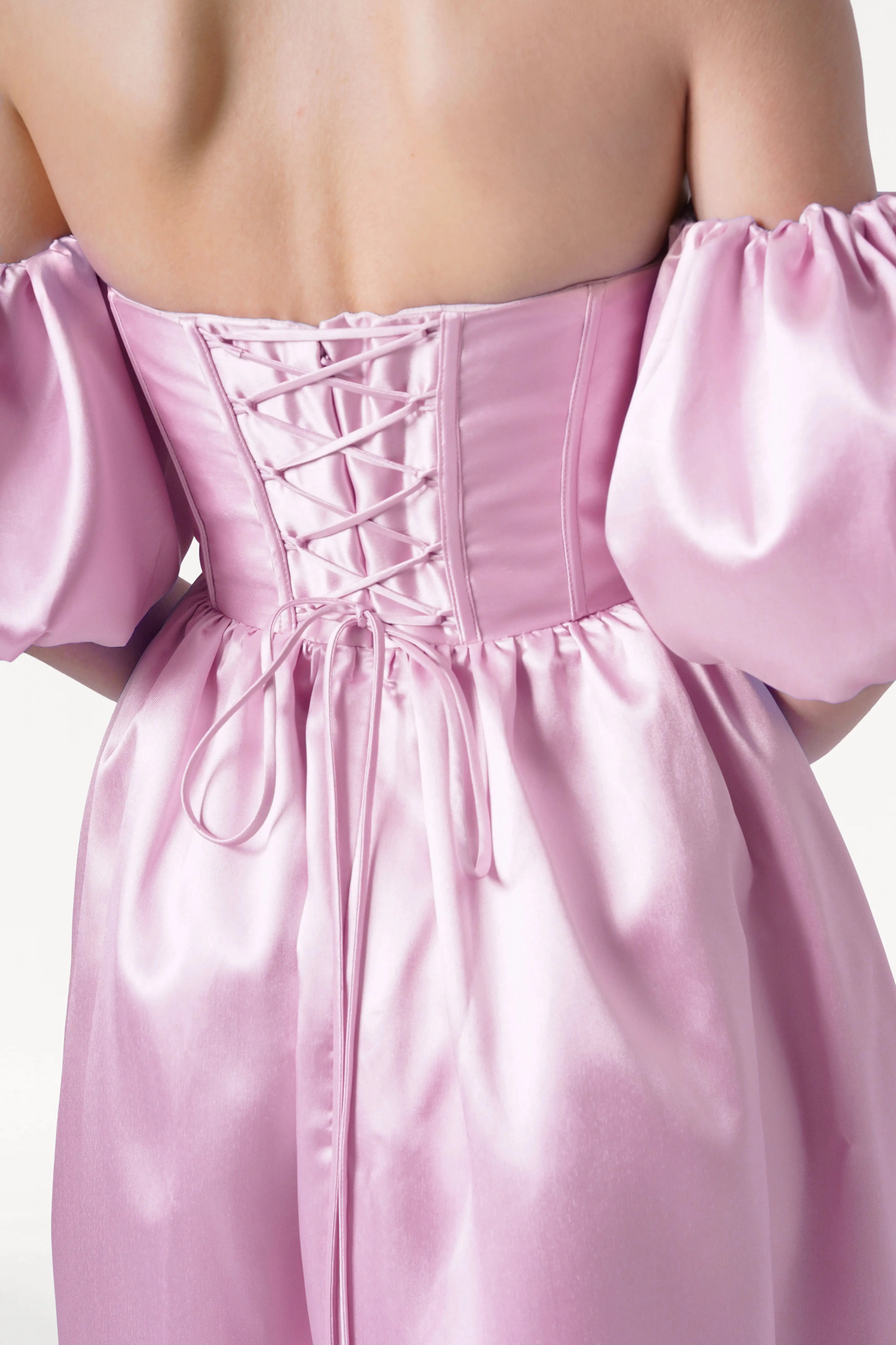 A person wearing the TH&TH Blair Bridesmaid Dress in Bubblegum Pink Satin Twill, an off-shoulder corset style with puffed detachable sleeves and a lace-up back, is shown from behind.