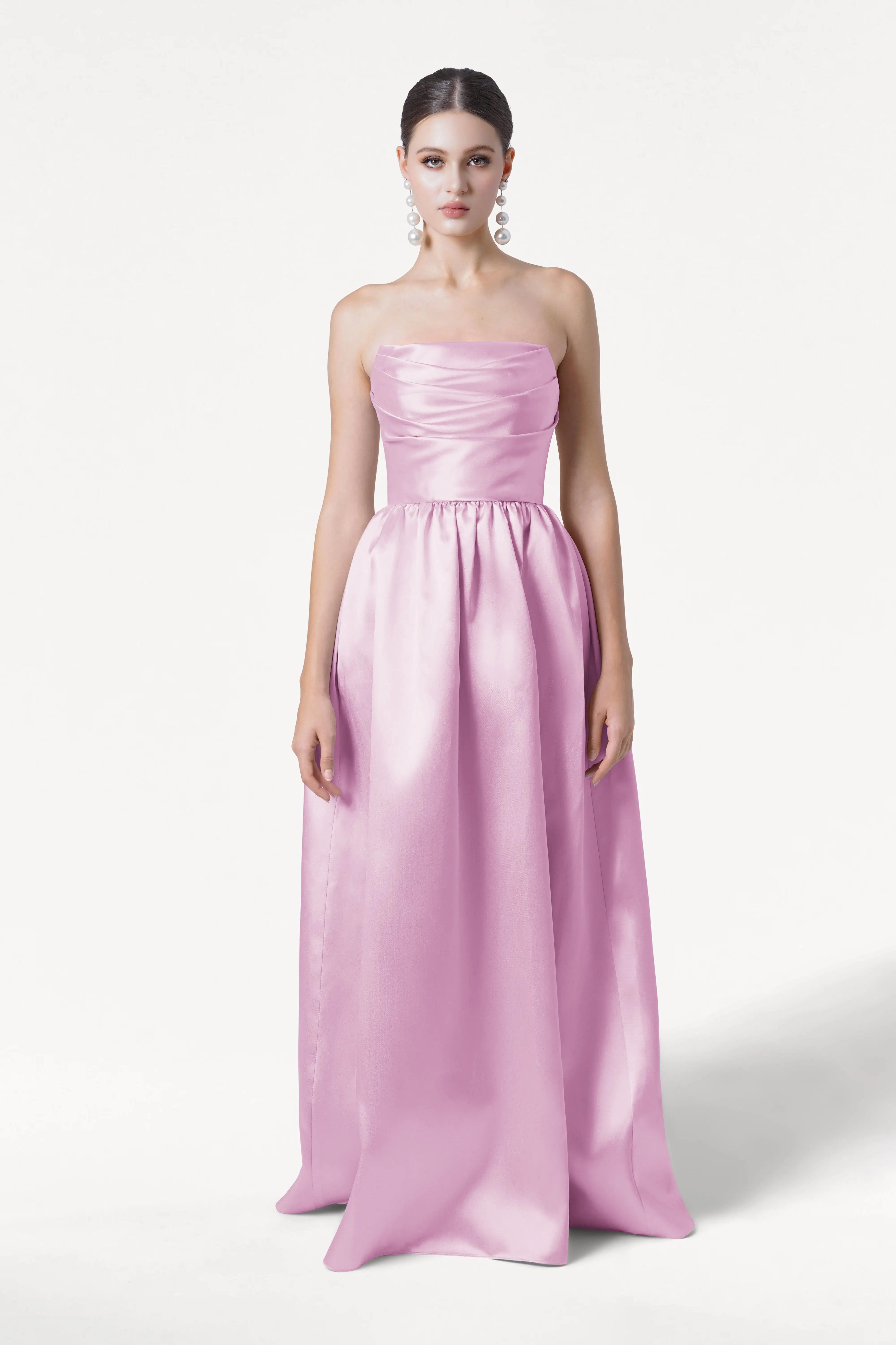 A woman faces forward in the TH&TH Blair Bridesmaid Dress in Bubblegum Pink Satin Twill, featuring a strapless, floor-length silhouette with a fitted bodice and full skirt. She has dark hair styled back and wears large pearl earrings against a white background.