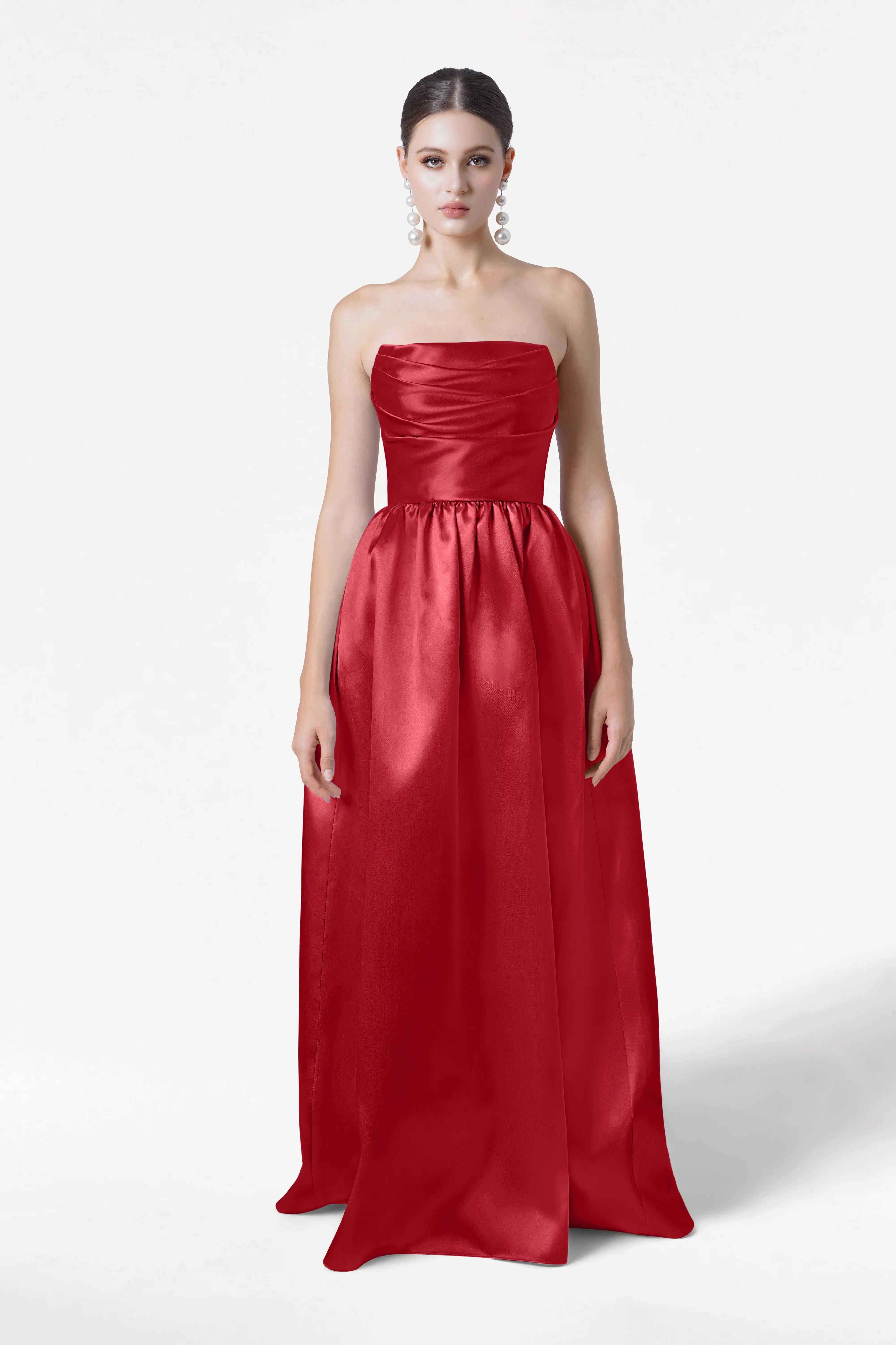 A woman stands against a white background wearing the Blair Bridesmaid Dress in Dark Cherry Satin Twill by TH&TH, which is strapless and floor-length with a fitted bodice and full skirt. Her hair is pulled back and she wears large dangling earrings.
