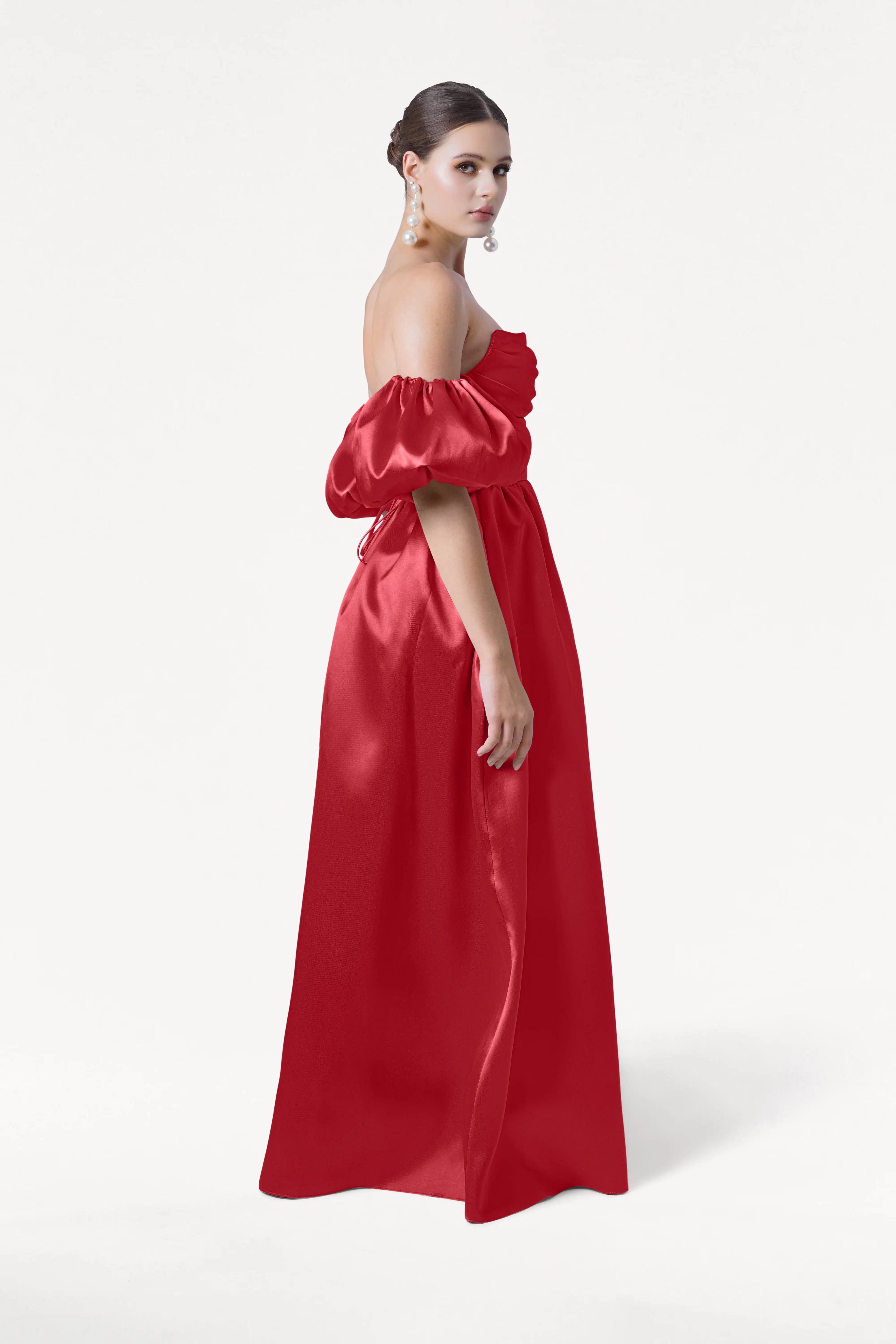 A woman wearing the TH&TH Blair Bridesmaid Dress in Dark Cherry Satin Twill with detachable puff sleeves stands sideways against a white background. Her hair is styled in a low bun and she accessorizes with long, dangling earrings.