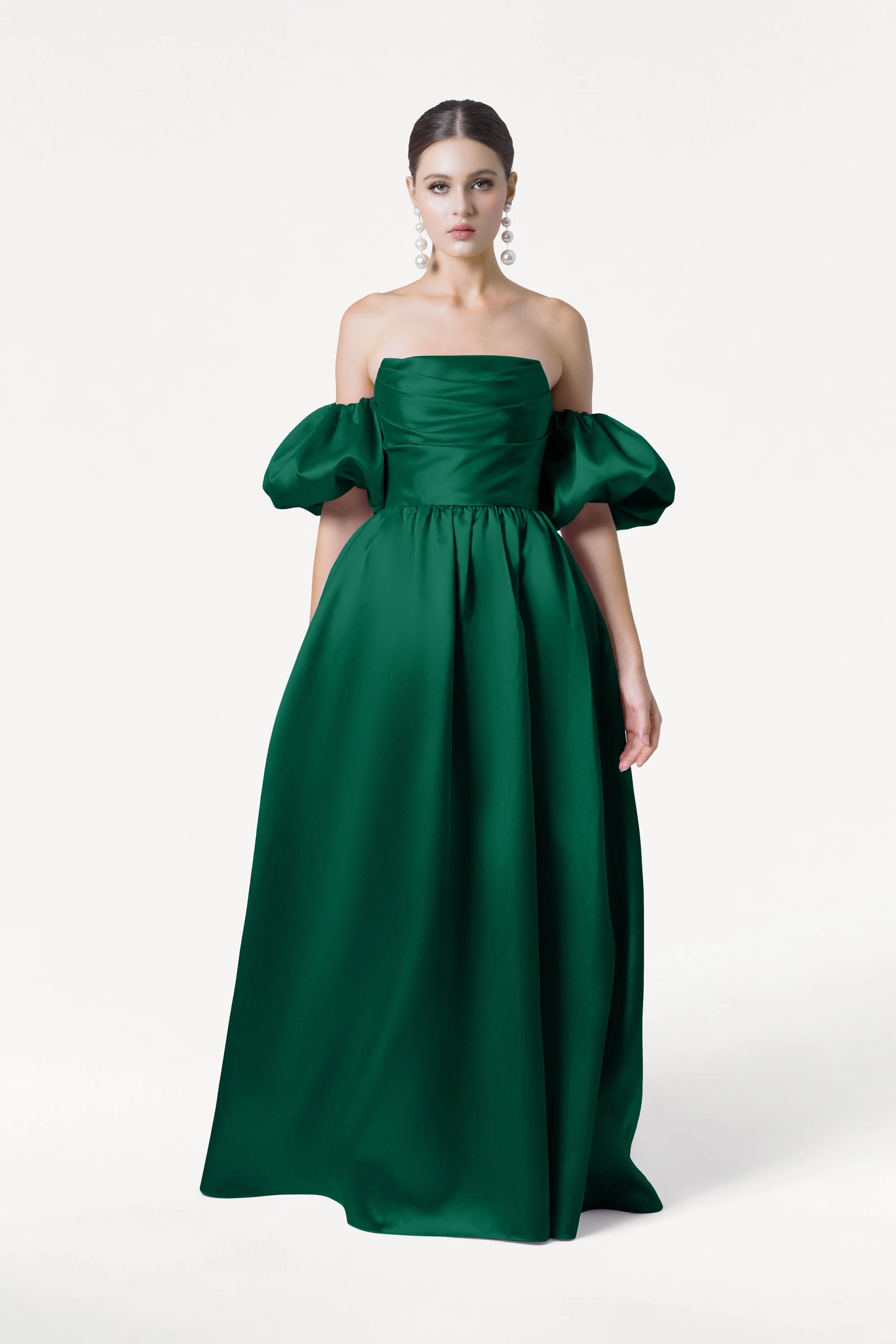 A woman stands against a white background in the TH&TH Blair Bridesmaid Dress in Forest Green Satin Twill, a floor-length gown with detachable puff sleeves. Her dark hair is pulled back, and she wears large dangling earrings.