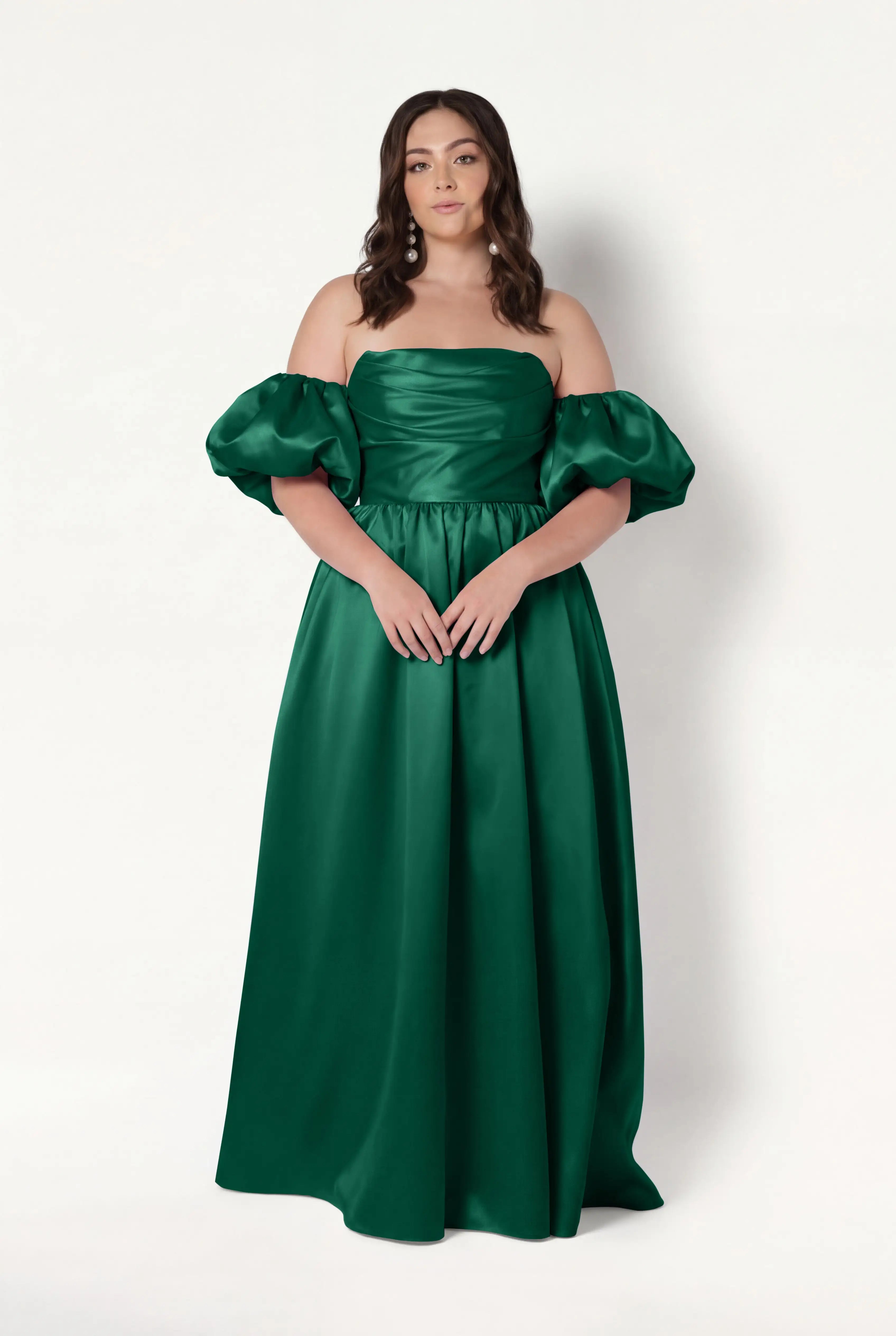 A woman with wavy brown hair and a neutral expression wears the TH&TH Blair Bridesmaid Dress in Forest Green Satin Twill, featuring an off-the-shoulder design, detachable puff sleeves, fitted bodice, and full floor-length skirt.