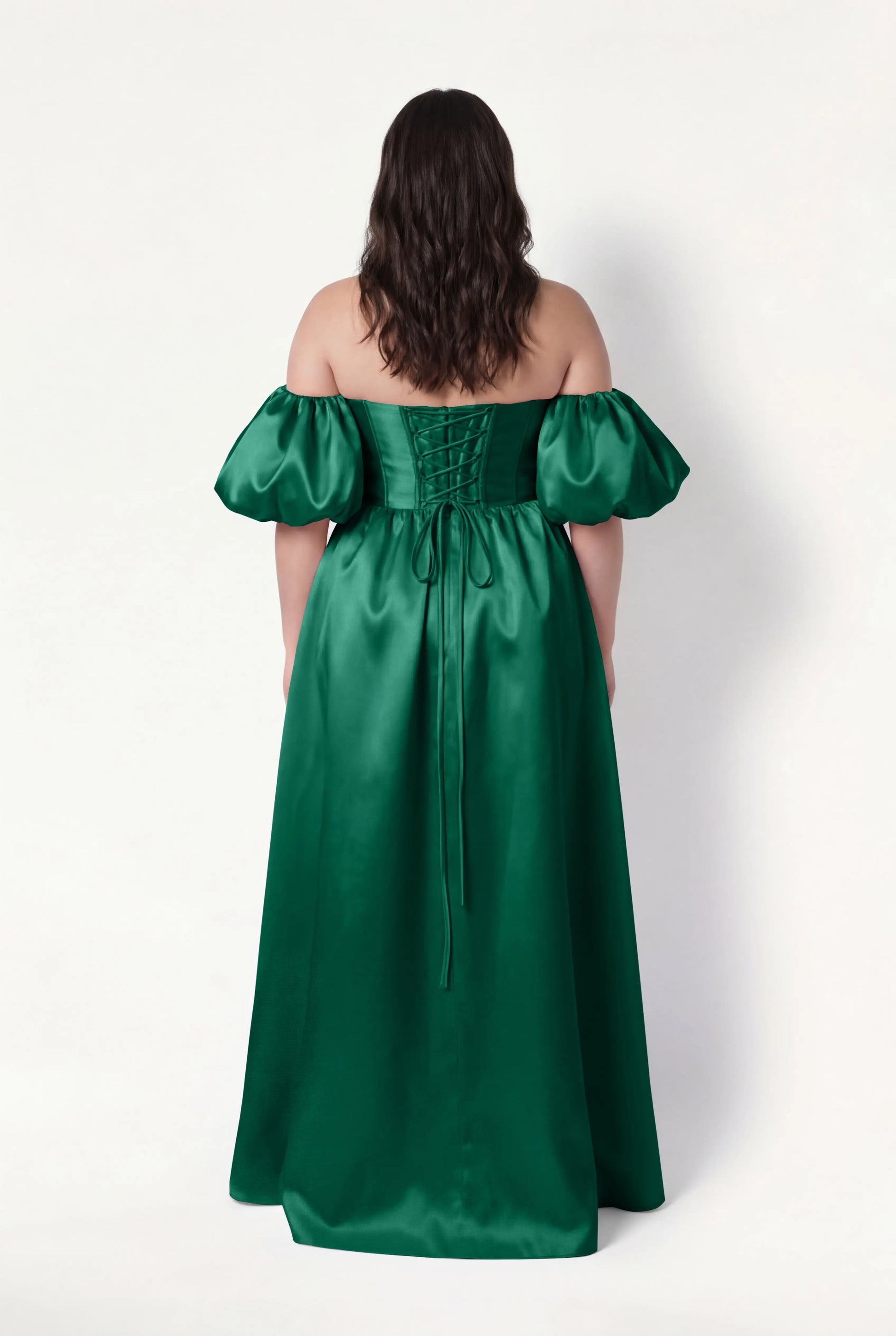 A woman with wavy brown hair models the TH&TH Blair Bridesmaid Dress in Forest Green Satin Twill, featuring an off-the-shoulder neckline, detachable puff sleeves, and a lace-up back, against a plain white background.