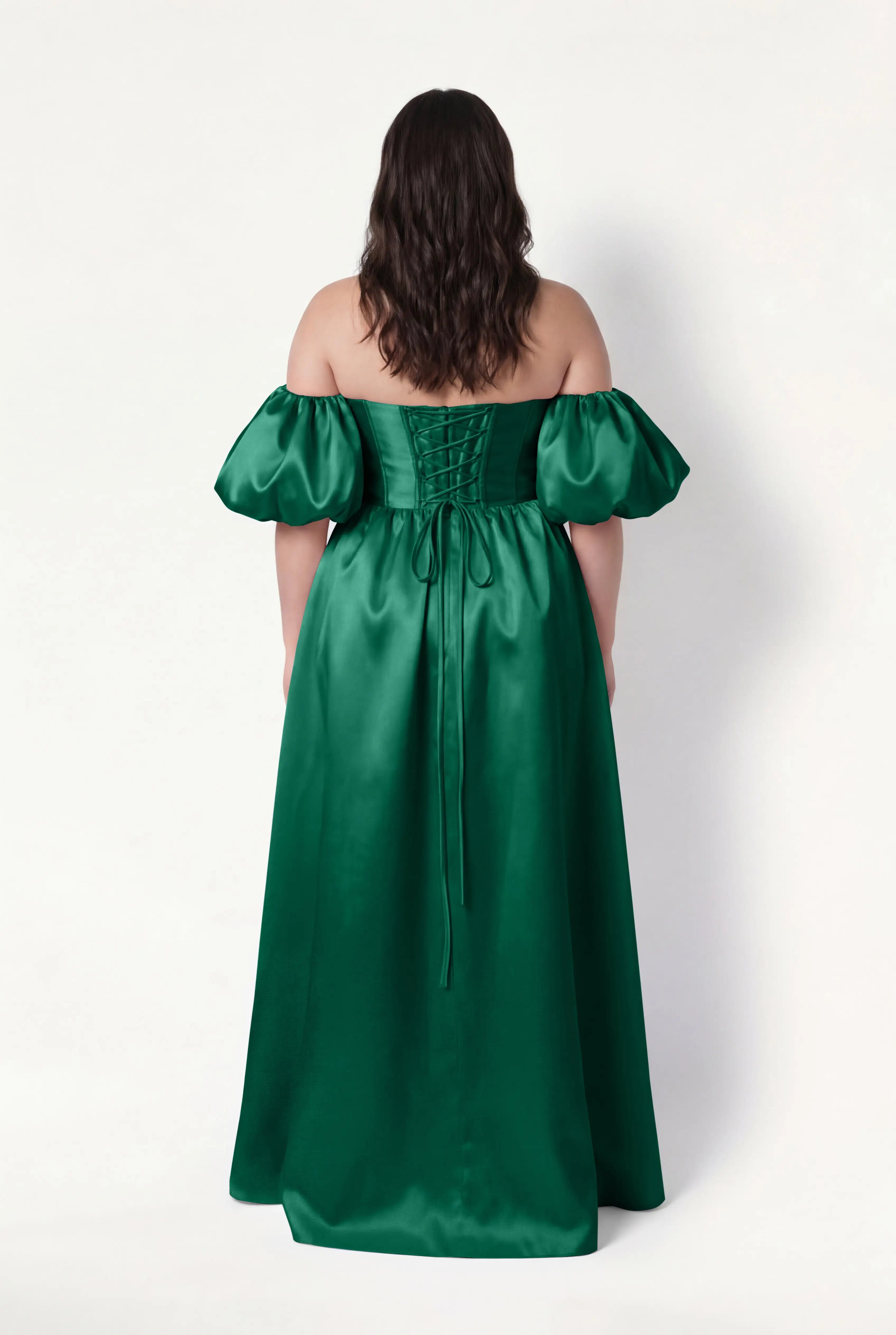 A woman with wavy brown hair models the TH&TH Blair Bridesmaid Dress in Forest Green Satin Twill, featuring an off-the-shoulder neckline, detachable puff sleeves, and a lace-up back, against a plain white background.