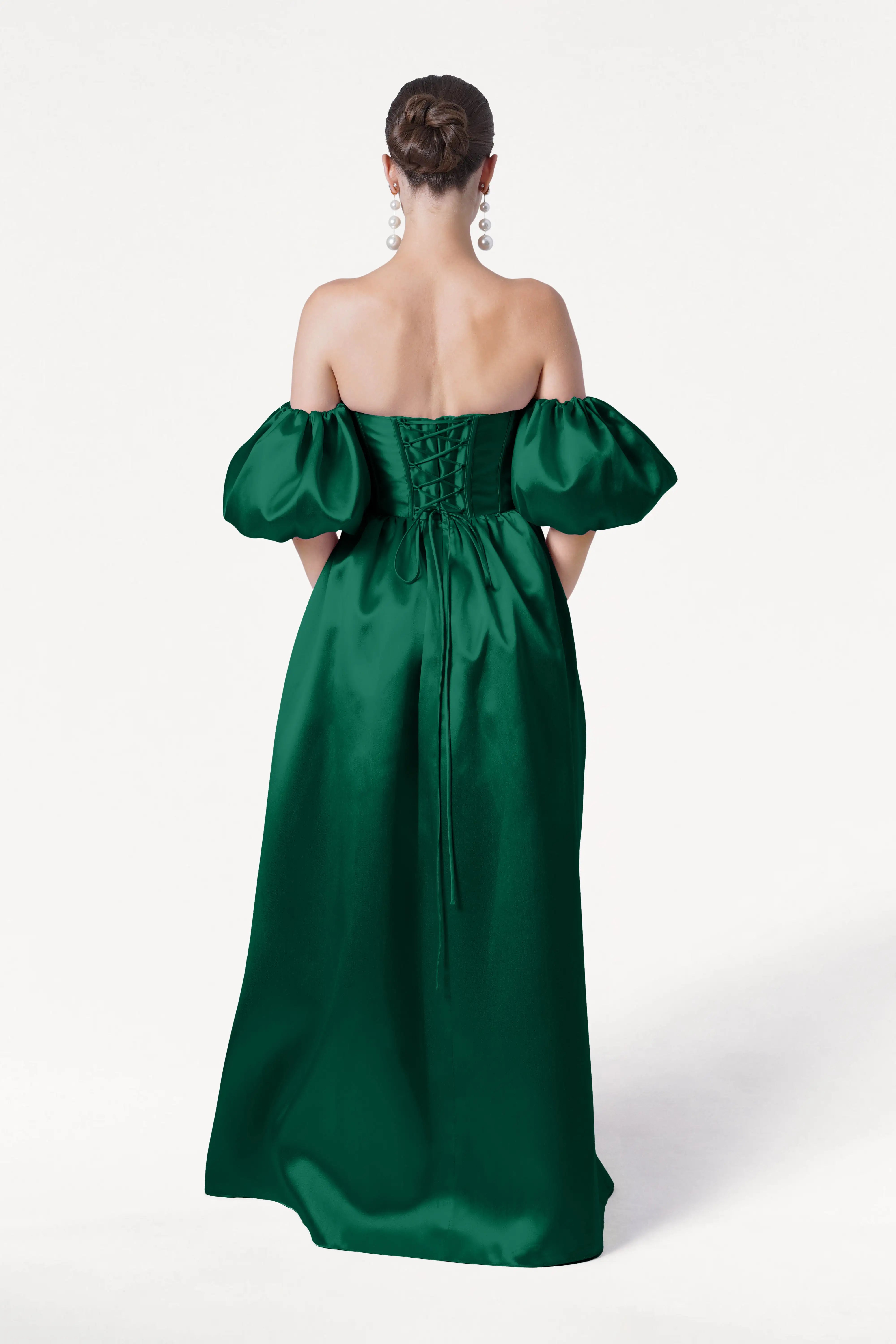 A woman faces away, wearing the TH&TH Blair Bridesmaid Dress in Forest Green Satin Twill with detachable puff sleeves and a lace-up back. Her hair is styled in an elegant bun, and she wears large dangling earrings against a plain white background.