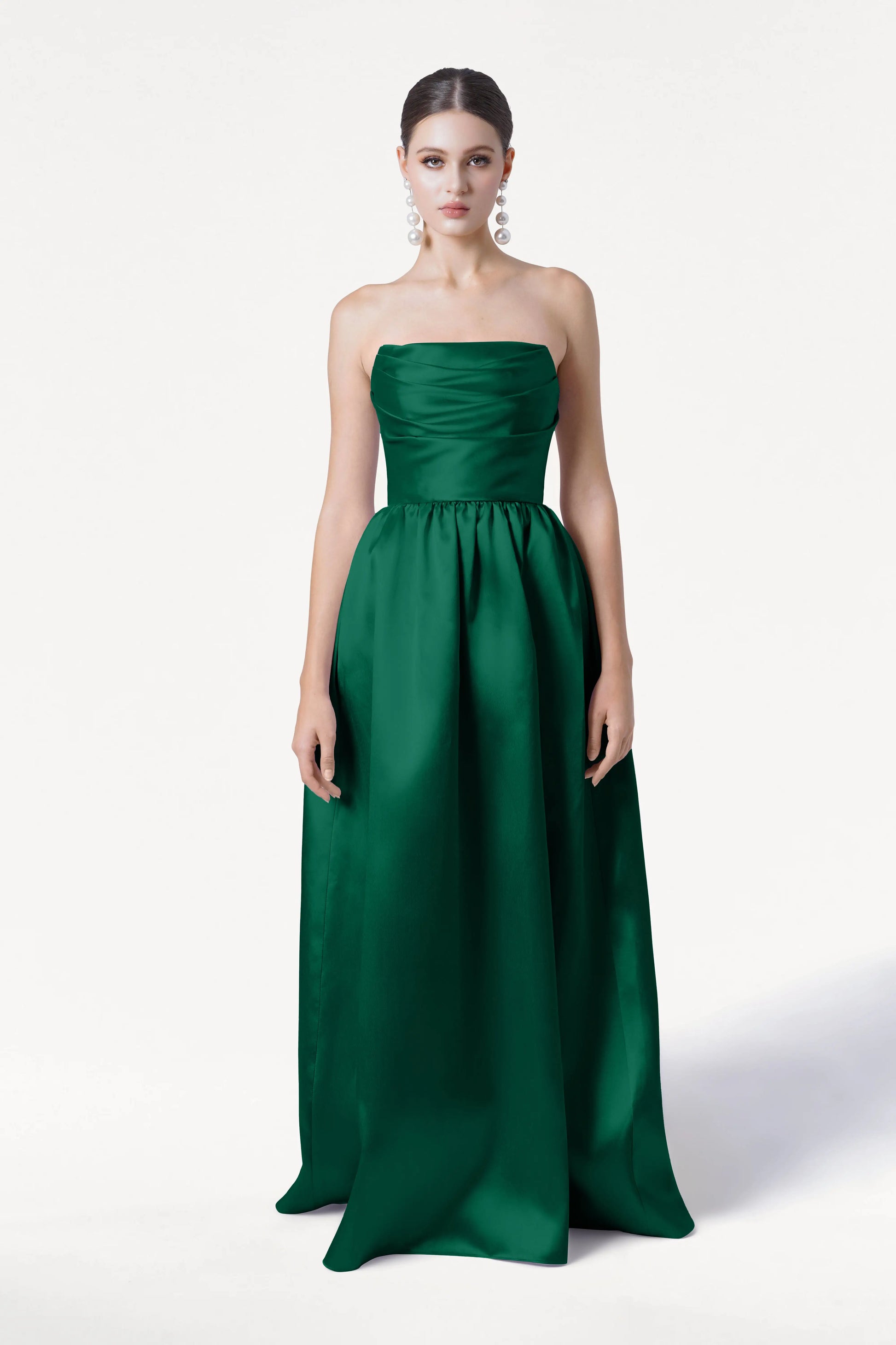A woman wears the TH&TH Blair Bridesmaid Dress in Forest Green Satin Twill, a strapless, floor-length gown with a fitted bodice, full skirt, and detachable puff sleeves. Her hair is styled back and she completes the look with large statement earrings.