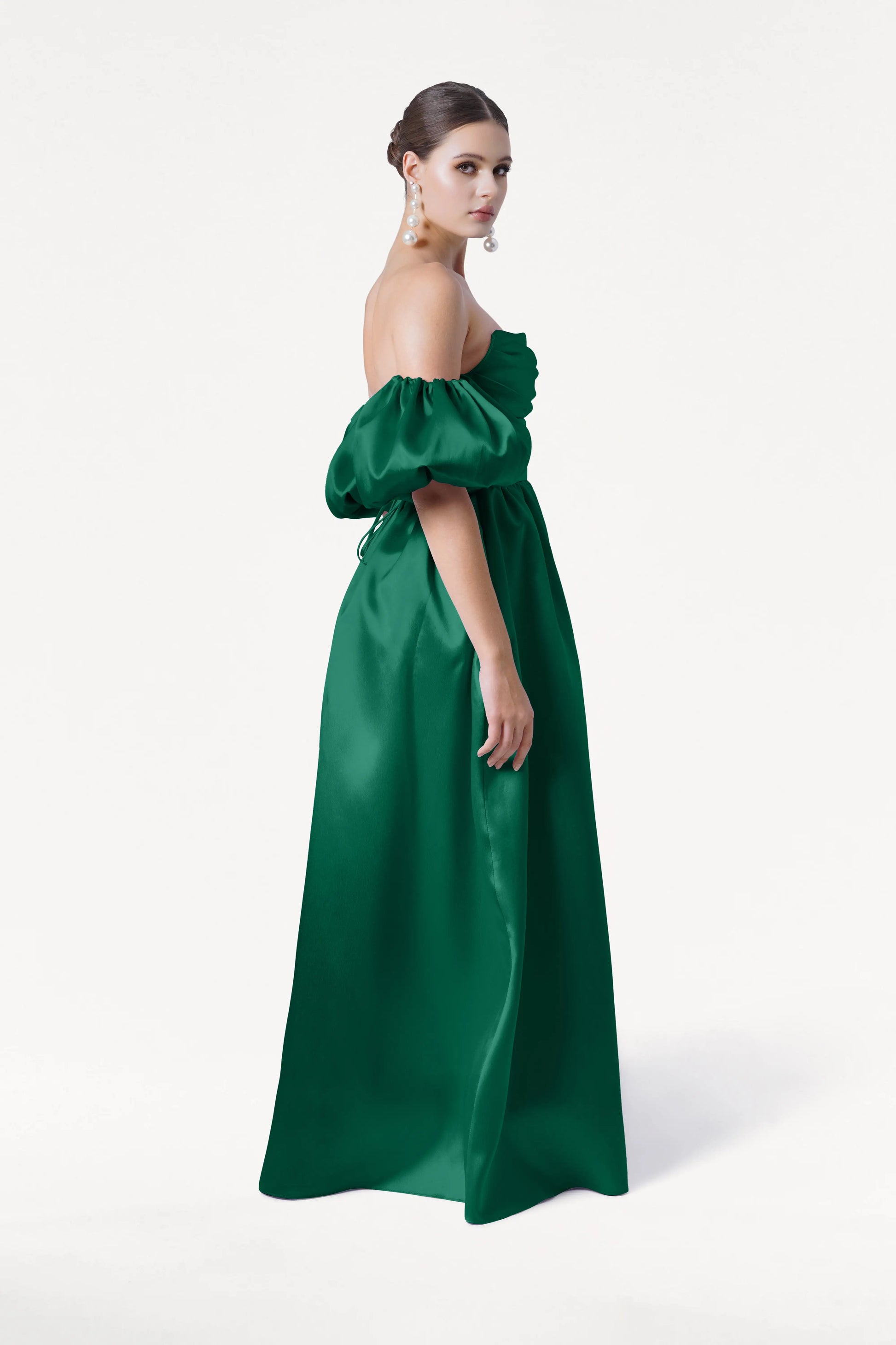 A woman models the TH&TH Blair Bridesmaid Dress in Forest Green Satin Twill, standing sideways against a white background. Her hair is pulled back, and she wears large dangling earrings.