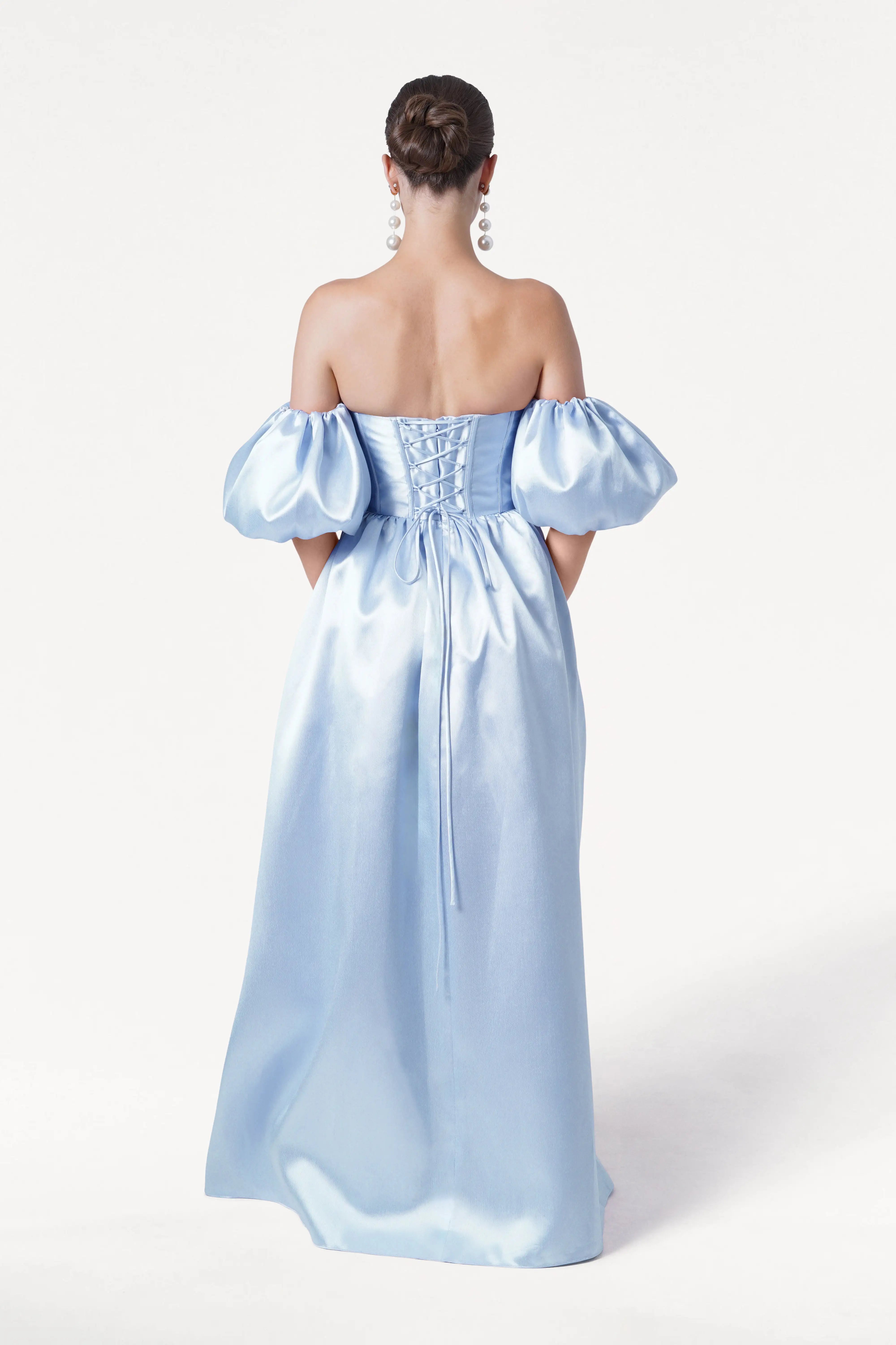 A woman faces away wearing the Blair Bridesmaid Dress in Frosted Blue Satin Twill by TH&TH. The shiny gown features detachable puff sleeves, an updo hairstyle, and large earrings set against a plain white background.